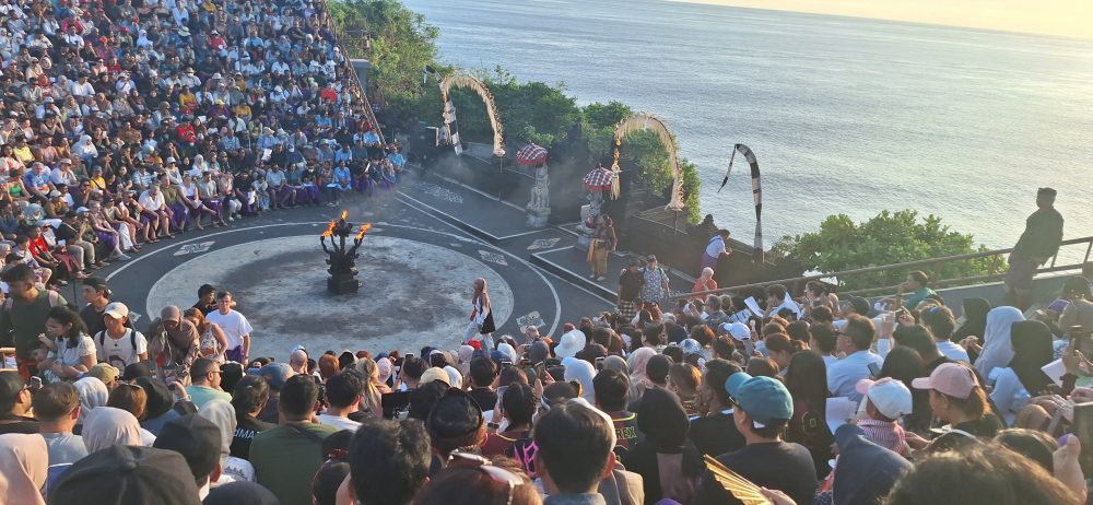 Sejarah Tari Kecak Bali: Pertemuan Timur dan Barat Sejarah Tari Kecak tidak bermula di panggung pertunjukan wisata, melainkan berakar jauh pada ritual keagamaan kuno yang disebut Sanghyang. Sanghyang merupakan tradisi tarian trance yang dilakukan untuk mengusir roh jahat, memohon perlindungan dari dewa, atau menyucikan komunitas dari wabah penyakit. Dalam ritual aslinya, para penari berada dalam kondisi tidak sadar dan diyakini dapat berkomunikasi dengan roh leluhur atau dewa-dewa. Paduan suara pria yang mengiringi ritual ini dikenal sebagai "Cak," yang berfungsi untuk menjaga ritme dan menciptakan atmosfer sakral yang intens. Transformasi dari ritual penyembuhan menuju seni pertunjukan dramatis terjadi pada tahun 1930-an, sebuah periode krusial dalam sejarah seni rupa dan pertunjukan Bali. Inovasi ini merupakan hasil kolaborasi antara I Wayan Limbak, seorang penari dan seniman visioner asal Bali, dengan Walter Spies, seorang pelukis berkebangsaan Jerman yang memiliki keterikatan emosional mendalam dengan budaya Bali. Spies, yang juga memiliki latar belakang dalam dunia musik dan perfilman, melihat potensi artistik yang luar biasa dalam paduan suara Sanghyang. Bersama Limbak, ia merancang sebuah koreografi yang menggabungkan elemen paduan suara "Cak" dengan narasi epos Ramayana. Evolusi ini terdokumentasi dengan baik dalam sejarah perkembangan genre tari Bali. Sebelum tahun 1930-an, elemen "Cak" hanyalah bagian dari ritus pengusiran setan Sanghyang Dedari. Namun, visi Spies dan Limbak di Desa Bona, Kabupaten Gianyar, berhasil mengubah paduan suara tersebut menjadi media penceritaan dramatis. Salah satu katalisator utama transformasi ini adalah keterlibatan Walter Spies dalam pembuatan film Insel der Dämonen (Pulau Iblis) pada tahun 1933 oleh Victor von Plessen. Spies menggunakan medium film untuk mendokumentasikan transformasi genre ini, yang pada gilirannya memperkenalkan estetika Kecak ke audiens internasional. Evolusi dan Pencapaian Historis Tari Kecak Periode/Tahun Milestone Sejarah Keterangan Signifikan Pra-1930 Era Ritual Sanghyang Berfungsi sebagai ritual sakral penyucian dan penolak bala. 1930-an Kolaborasi Limbak-Spies Penciptaan format modern di Desa Bona, Gianyar; integrasi kisah Ramayana. 1933 Dokumentasi Sinematik Penayangan film Insel der Dämonen yang memuat elemen visual Kecak. 1970-an Ekspansi Regional Kecak mulai dipentaskan di luar Gianyar, termasuk di sekolah-sekolah seni di Bali. 1979 Rekor Massal Pertama Pementasan dengan 500 penari pria yang membawakan epos Mahabharata. 2006 Rekor Dunia Tabanan Pementasan kolosal 5.000 penari di Tanah Lot untuk mendongkrak pariwisata. Anatomi Suara: Orkestrasi Gamelan Manusia Keunikan yang paling mencolok dari Tari Kecak adalah absennya instrumen gamelan logam yang biasanya menjadi tulang punggung musik tradisional Bali. Dalam Kecak, instrumen musiknya adalah suara manusia itu sendiri. Orkestrasi vokal ini begitu kompleks sehingga mampu menghasilkan berbagai macam tekstur bunyi, mulai dari dentuman ritmis yang cepat hingga melodi yang melankolis. Paduan suara pria yang duduk melingkar, yang disebut sebagai pengecak, tidak semata-mata meneriakkan kata "cak" secara acak. Terdapat pembagian peran yang sangat teknis di dalam lingkaran tersebut untuk menjaga keharmonisan dan dinamika pertunjukan. Ritme ini dipercaya memiliki frekuensi yang dapat memengaruhi kesadaran, menciptakan suasana magis yang memungkinkan para penari dan penonton terhanyut dalam narasi. Satu-satunya instrumen fisik yang digunakan adalah kincringan, yaitu lonceng kecil yang diikatkan pada kaki para penari lakon (Rama, Sita, Hanuman) untuk memberikan aksen pada setiap langkah dan gerakan tari klasik mereka. Geometri Spiritual: Formasi Lingkaran dan Keseimbangan Rwa Bhineda Visualisasi Tari Kecak dibangun di atas simbolisme yang sarat makna. Formasi para penari yang duduk melingkar bukan sekadar pilihan estetika ruang, melainkan representasi dari kesatuan kosmos dan harmoni alam semesta. Dalam pandangan Hindu Bali, lingkaran melambangkan siklus kehidupan yang abadi, tanpa awal dan tanpa akhir. Posisi ini juga mencerminkan nilai-nilai sosial masyarakat Bali yang menekankan pada gotong royong, kebersamaan, dan kesetaraan; setiap penari berkontribusi pada energi kolektif yang kuat tanpa ada individu yang berusaha menonjolkan diri secara berlebihan. Di tengah lingkaran tersebut, biasanya terdapat damar kecak atau obor api yang berfungsi sebagai titik pusat energi. Api dalam budaya Bali memiliki fungsi ganda: sebagai penerang dan sebagai penyuci (Agni). Menariknya, penggunaan lampu tengah ini awalnya merupakan inovasi teknis yang diperkenalkan oleh Walter Spies untuk kebutuhan pencahayaan saat proses syuting film, agar wajah para penari dapat tertangkap kamera dengan lebih jelas. Namun, seiring berjalannya waktu, elemen ini menjadi bagian permanen dan esensial dalam setiap pertunjukan Kecak, menambah nuansa dramatis dan sakral. Elemen simbolis lainnya adalah kostum kain poleng yang dikenakan oleh para pengecak. Kain dengan motif kotak-kotak hitam dan putih ini merupakan manifestasi dari filosofi Rwa Bhineda, sebuah ajaran fundamental tentang dualisme yang harmonis. Warna hitam melambangkan kegelapan, malam, atau aspek negatif kehidupan, sementara warna putih melambangkan cahaya, siang, atau aspek positif. Dalam pandangan masyarakat Bali, kedua kekuatan ini tidak dapat dipisahkan dan harus selalu berada dalam keseimbangan untuk menjaga keharmonisan dunia. Filosofi ini mengajarkan bahwa kehidupan tidak pernah bersifat absolut; ada keindahan di dalam kegelapan dan ada tantangan di dalam cahaya. Cerita Ramayana dalam Tari Kecak Meskipun instrumen vokalnya berasal dari ritual Sanghyang, jiwa cerita Tari Kecak adalah epos Ramayana. Pertunjukan biasanya menampilkan cuplikan paling dramatis dari perjalanan Pangeran Rama, yang melambangkan pertarungan abadi antara kebajikan (Dharma) dan kejahatan (Adharma). Narasi dimulai di Hutan Dandaka, tempat Rama, istrinya Dewi Sita, dan adiknya Laksmana menjalani masa pengasingan. Di sini, penonton disuguhi drama penculikan yang penuh tipu muslihat. Rahwana, raja raksasa dari Kerajaan Alengka yang dikuasai nafsu serakah, mengirim anak buahnya yang menyamar sebagai kijang emas untuk memikat hati Sita. Ketertarikan Sita pada kijang tersebut memaksa Rama pergi berburu, meninggalkan Sita di bawah perlindungan Laksmana. Adegan yang seringkali menyentuh hati penonton adalah pengorbanan Burung Garuda, Jatayu, yang mencoba mengadang Rahwana saat membawa terbang Sita ke Alengka. Meskipun Jatayu harus gugur setelah sayapnya ditebas oleh kesaktian Rahwana, tindakannya menjadi simbol moral tentang keberanian dan pengorbanan demi membela kehormatan. Puncak ketegangan terjadi saat kemunculan Hanoman, sang kera putih sakti yang menjadi utusan Rama. Hanoman bukan hanya karakter heroik, tetapi juga seringkali menjadi pencair suasana melalui gerakan-gerakannya yang lincah dan jenaka, bahkan terkadang berinteraksi langsung dengan penonton. Adegan "Hanoman Obong" atau pembakaran Hanoman di Alengka adalah momen yang paling ditunggu-tunggu. Di atas bara api yang menyala, penari Hanoman menari dengan tangkas, menyebarkan api ke segala penjuru istana Rahwana, sebuah simbolisasi dari kemurnian yang tidak dapat hangus oleh api kejahatan. Setiap gerakan dalam tarian ini—mulai dari gerakan tangan (mudra), ekspresi wajah (abhinaya), hingga langkah kaki yang anggun—memiliki makna tersendiri dalam teknik tari Bali klasik. Kesetiaan Sita kepada suaminya, keteguhan hati Rama, dan pengabdian Hanoman memberikan pesan moral yang mendalam bagi para penonton: bahwa kebenaran mungkin mengalami hambatan, namun pada akhirnya akan selalu menemukan jalannya untuk menang. So, tokoh-tokoh utama pun hadir: Rama, sang pangeran Sinta, istrinya Hanoman, kera putih yang setia Rahwana, raja raksasa Pertunjukan Tari Kecak biasanya mengangkat kisah Ramayana, yang berfokus pada perjuangan Rama menyelamatkan Sinta dari penculikan Rahwana. Dalam perjalanan tersebut, Rama dibantu oleh Hanoman, tokoh kera putih yang menjadi salah satu karakter paling ikonik dalam pertunjukan ini. Adegan Hanoman bermain dengan api biasanya menjadi bagian paling ditunggu oleh penonton. Yang membuatnya istimewa bukan hanya ceritanya, tetapi cara penyampaiannya. Tanpa dialog panjang, tanpa efek suara modern, semua emosi disampaikan melalui gerakan, ekspresi, dan tentu saja—suara “cak” yang terus mengalun. Pada puncak pertunjukan, adegan api sering kali menjadi klimaks. Api menyala, Hanoman menari di sekitarnya, dan suasana berubah menjadi intens sekaligus memukau. Lokasi Terbaik Menonton Tari Kecak di Bali Berikut beberapa tempat terbaik untuk menyaksikan Tari Kecak Bali: 1. Pura Luhur Uluwatu Lokasi paling populer dengan latar tebing dan sunset langsung ke Samudra Hindia. Suasana sangat dramatis. 2. Pura Dalem Ubud Cocok untuk yang menginap di Ubud. Suasana lebih intim dan kental nuansa budaya. 3. Garuda Wisnu Kencana Area luas dengan fasilitas modern, cocok untuk wisata keluarga. Harga Tiket Tari Kecak Bali Harga tiket menonton Tari Kecak cukup bervariasi tergantung lokasi: Uluwatu: Rp150.000 – Rp200.000 Ubud: Rp100.000 – Rp150.000 GWK: mulai Rp100.000 Harga bisa berubah sewaktu-waktu, terutama saat musim liburan. Jam Pertunjukan Tari Kecak Umumnya, Tari Kecak ditampilkan pada: Pukul 18.00 – 19.00 WITA Durasi: sekitar 60 menit Waktu ini dipilih karena bertepatan dengan sunset, sehingga menciptakan suasana yang lebih dramatis. Betewe, karena pertunjukan ini sangat populer, disarankan untuk datang lebih awal agar mendapatkan tempat duduk terbaik—terutama di lokasi seperti Uluwatu yang sering penuh. DIantara tiga destinasi di atas, saya masih berkunjung yang di Uluwatu. Sore itu, begitu matahari mulai merunduk di cakrawala, semburat jingga keemasan yang membasuh tebing-tebing kapur di Pura Luhur Uluwatu bukan sekadar penanda berakhirnya hari, melainkan aba-aba bagi dimulainya sebuah upacara estetika yang telah memikat dunia selama hampir satu abad. Di sebuah amfiteater terbuka yang bertengger megah di atas Samudra Hindia, ratusan pria duduk melingkar dengan dada telanjang, hanya mengenakan kain poleng hitam-putih yang melilit pinggang mereka. Ketika matahari bersentuhan dengan garis laut, sebuah seruan ritmik memecah keheningan: "cak, cak, cak." Suara itu bukan sekadar iringan musik, melainkan sebuah getaran kolektif yang merambat dari bumi, melalui tubuh-tubuh penari, hingga menyentuh relung spiritual setiap penonton yang hadir. Awalnya pelan. Kemudian semakin kuat, berlapis, dan ritmis. Suara itu seperti gelombang yang datang silih berganti, membentuk harmoni yang tidak biasa. Inilah ciri khas utama Tari Kecak—tarian yang tidak menggunakan alat musik, melainkan mengandalkan vokal kolektif sebagai pengiring utama. Tari Kecak, sebuah mahakarya yang lahir dari rahim tradisi sakral dan visi artistik modern, berdiri sebagai monumen hidup bagi identitas budaya Bali yang tak pernah lekang oleh zaman. Pementasan ini merupakan perpaduan unik antara energi, cerita, dan spiritualitas yang membius. Berbeda dengan tarian Bali lainnya yang mengandalkan dentuman gamelan perunggu, Kecak sepenuhnya mengandalkan instrumen vokal manusia. Harmoni suara yang dihasilkan oleh 50 hingga 150 penari pria ini menciptakan lanskap bunyi yang trance-like, membawa penonton ke dalam dunia epos Ramayana yang penuh dengan pesan moral tentang kesetiaan, keberanian, dan kemenangan kebenaran atas kejahatan. Di balik kemegahan teatrikalnya, Kecak menyimpan lapisan sejarah yang dalam, mulai dari ritual penyucian kuno hingga inovasi seni di tahun 1930-an yang melibatkan seniman lokal dan pelukis mancanegara. Bagi banyak wisatawan, pengalaman ini terasa magis. Suara manusia yang berpadu dalam pola kompleks menciptakan suasana yang hampir transendental. Salah satu alasan mengapa Tari Kecak begitu berkesan adalah lokasi pertunjukannya. Di Pura Luhur Uluwatu, misalnya, panggung terbuka menghadap langsung ke Samudra Hindia. Tebing tinggi menjadi latar alami, sementara matahari terbenam menciptakan pencahayaan dramatis yang sulit ditandingi. Bagi sebagian orang, Tari Kecak mungkin terlihat seperti atraksi wisata. Namun bagi masyarakat Bali, ini adalah bagian dari identitas. Suara “cak” yang berulang bukan sekadar irama, melainkan simbol kebersamaan. Puluhan bahkan ratusan penari bergerak dan bersuara sebagai satu kesatuan—tanpa dominasi individu. Di tengah dunia yang semakin individualistik, pesan ini terasa relevan. Tari Kecak mengajarkan bahwa harmoni tercipta ketika setiap elemen bekerja bersama. Tips Menikmati Tari Kecak Agar pengalaman semakin maksimal, berikut beberapa tips: Datang minimal 30–60 menit sebelum pertunjukan Pilih tempat duduk dengan sudut pandang sunset Gunakan pakaian nyaman (karena duduk cukup lama) Bawa kamera, tapi jangan lupa menikmati momen tanpa layar Dan yang terpenting, datanglah dengan pikiran terbuka. Karena Tari Kecak bukan hanya untuk ditonton, tetapi untuk dirasakan. Harmoni dalam Setiap Suara Saat pertunjukan berakhir dan suara “cak” perlahan mereda, suasana terasa hening. Penonton berdiri, memberikan tepuk tangan panjang, seolah tidak ingin momen itu berakhir. Di langit, matahari telah tenggelam. Namun pengalaman itu tetap tinggal. Tari Kecak adalah bukti bahwa budaya tidak pernah benar-benar diam. Ia hidup, bergerak, dan terus beradaptasi. Di tengah modernitas Bali yang terus berkembang, Tari Kecak tetap menjadi pengingat bahwa akar budaya adalah kekuatan yang menjaga identitas. Tari Kecak bukan sekadar tontonan, melainkan sebuah simfoni kehidupan. Di dalamnya terdengar suara tanah Bali, getaran doa para leluhur, dan semangat kebersamaan yang terus hidup melampaui batas waktu. Ketika suara "cak" terakhir menggema dan api di tengah lingkaran perlahan padam, yang tersisa bukan hanya kenangan akan sebuah pertunjukan, melainkan sebuah pemahaman yang lebih dalam tentang harmoni manusia dengan sesama, alam, dan Tuhan. Bagi siapa pun yang pernah menyaksikannya, Kecak adalah detak jantung Bali yang akan terus berdenyut, mengundang dunia untuk merayakan keindahan dalam kesatuan vokal dan spiritualitas yang tak tergantikan.