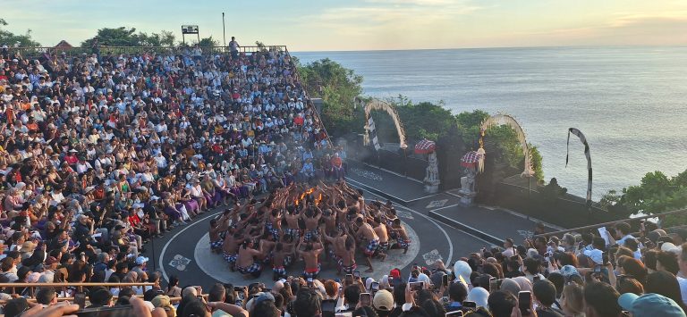 Sejarah Tari Kecak Bali: Pertemuan Timur dan Barat Sejarah Tari Kecak tidak bermula di panggung pertunjukan wisata, melainkan berakar jauh pada ritual keagamaan kuno yang disebut Sanghyang. Sanghyang merupakan tradisi tarian trance yang dilakukan untuk mengusir roh jahat, memohon perlindungan dari dewa, atau menyucikan komunitas dari wabah penyakit. Dalam ritual aslinya, para penari berada dalam kondisi tidak sadar dan diyakini dapat berkomunikasi dengan roh leluhur atau dewa-dewa. Paduan suara pria yang mengiringi ritual ini dikenal sebagai "Cak," yang berfungsi untuk menjaga ritme dan menciptakan atmosfer sakral yang intens. Transformasi dari ritual penyembuhan menuju seni pertunjukan dramatis terjadi pada tahun 1930-an, sebuah periode krusial dalam sejarah seni rupa dan pertunjukan Bali. Inovasi ini merupakan hasil kolaborasi antara I Wayan Limbak, seorang penari dan seniman visioner asal Bali, dengan Walter Spies, seorang pelukis berkebangsaan Jerman yang memiliki keterikatan emosional mendalam dengan budaya Bali. Spies, yang juga memiliki latar belakang dalam dunia musik dan perfilman, melihat potensi artistik yang luar biasa dalam paduan suara Sanghyang. Bersama Limbak, ia merancang sebuah koreografi yang menggabungkan elemen paduan suara "Cak" dengan narasi epos Ramayana. Evolusi ini terdokumentasi dengan baik dalam sejarah perkembangan genre tari Bali. Sebelum tahun 1930-an, elemen "Cak" hanyalah bagian dari ritus pengusiran setan Sanghyang Dedari. Namun, visi Spies dan Limbak di Desa Bona, Kabupaten Gianyar, berhasil mengubah paduan suara tersebut menjadi media penceritaan dramatis. Salah satu katalisator utama transformasi ini adalah keterlibatan Walter Spies dalam pembuatan film Insel der Dämonen (Pulau Iblis) pada tahun 1933 oleh Victor von Plessen. Spies menggunakan medium film untuk mendokumentasikan transformasi genre ini, yang pada gilirannya memperkenalkan estetika Kecak ke audiens internasional. Evolusi dan Pencapaian Historis Tari Kecak Periode/Tahun Milestone Sejarah Keterangan Signifikan Pra-1930 Era Ritual Sanghyang Berfungsi sebagai ritual sakral penyucian dan penolak bala. 1930-an Kolaborasi Limbak-Spies Penciptaan format modern di Desa Bona, Gianyar; integrasi kisah Ramayana. 1933 Dokumentasi Sinematik Penayangan film Insel der Dämonen yang memuat elemen visual Kecak. 1970-an Ekspansi Regional Kecak mulai dipentaskan di luar Gianyar, termasuk di sekolah-sekolah seni di Bali. 1979 Rekor Massal Pertama Pementasan dengan 500 penari pria yang membawakan epos Mahabharata. 2006 Rekor Dunia Tabanan Pementasan kolosal 5.000 penari di Tanah Lot untuk mendongkrak pariwisata. Anatomi Suara: Orkestrasi Gamelan Manusia Keunikan yang paling mencolok dari Tari Kecak adalah absennya instrumen gamelan logam yang biasanya menjadi tulang punggung musik tradisional Bali. Dalam Kecak, instrumen musiknya adalah suara manusia itu sendiri. Orkestrasi vokal ini begitu kompleks sehingga mampu menghasilkan berbagai macam tekstur bunyi, mulai dari dentuman ritmis yang cepat hingga melodi yang melankolis. Paduan suara pria yang duduk melingkar, yang disebut sebagai pengecak, tidak semata-mata meneriakkan kata "cak" secara acak. Terdapat pembagian peran yang sangat teknis di dalam lingkaran tersebut untuk menjaga keharmonisan dan dinamika pertunjukan. Ritme ini dipercaya memiliki frekuensi yang dapat memengaruhi kesadaran, menciptakan suasana magis yang memungkinkan para penari dan penonton terhanyut dalam narasi. Satu-satunya instrumen fisik yang digunakan adalah kincringan, yaitu lonceng kecil yang diikatkan pada kaki para penari lakon (Rama, Sita, Hanuman) untuk memberikan aksen pada setiap langkah dan gerakan tari klasik mereka. Geometri Spiritual: Formasi Lingkaran dan Keseimbangan Rwa Bhineda Visualisasi Tari Kecak dibangun di atas simbolisme yang sarat makna. Formasi para penari yang duduk melingkar bukan sekadar pilihan estetika ruang, melainkan representasi dari kesatuan kosmos dan harmoni alam semesta. Dalam pandangan Hindu Bali, lingkaran melambangkan siklus kehidupan yang abadi, tanpa awal dan tanpa akhir. Posisi ini juga mencerminkan nilai-nilai sosial masyarakat Bali yang menekankan pada gotong royong, kebersamaan, dan kesetaraan; setiap penari berkontribusi pada energi kolektif yang kuat tanpa ada individu yang berusaha menonjolkan diri secara berlebihan. Di tengah lingkaran tersebut, biasanya terdapat damar kecak atau obor api yang berfungsi sebagai titik pusat energi. Api dalam budaya Bali memiliki fungsi ganda: sebagai penerang dan sebagai penyuci (Agni). Menariknya, penggunaan lampu tengah ini awalnya merupakan inovasi teknis yang diperkenalkan oleh Walter Spies untuk kebutuhan pencahayaan saat proses syuting film, agar wajah para penari dapat tertangkap kamera dengan lebih jelas. Namun, seiring berjalannya waktu, elemen ini menjadi bagian permanen dan esensial dalam setiap pertunjukan Kecak, menambah nuansa dramatis dan sakral. Elemen simbolis lainnya adalah kostum kain poleng yang dikenakan oleh para pengecak. Kain dengan motif kotak-kotak hitam dan putih ini merupakan manifestasi dari filosofi Rwa Bhineda, sebuah ajaran fundamental tentang dualisme yang harmonis. Warna hitam melambangkan kegelapan, malam, atau aspek negatif kehidupan, sementara warna putih melambangkan cahaya, siang, atau aspek positif. Dalam pandangan masyarakat Bali, kedua kekuatan ini tidak dapat dipisahkan dan harus selalu berada dalam keseimbangan untuk menjaga keharmonisan dunia. Filosofi ini mengajarkan bahwa kehidupan tidak pernah bersifat absolut; ada keindahan di dalam kegelapan dan ada tantangan di dalam cahaya. Cerita Ramayana dalam Tari Kecak Meskipun instrumen vokalnya berasal dari ritual Sanghyang, jiwa cerita Tari Kecak adalah epos Ramayana. Pertunjukan biasanya menampilkan cuplikan paling dramatis dari perjalanan Pangeran Rama, yang melambangkan pertarungan abadi antara kebajikan (Dharma) dan kejahatan (Adharma). Narasi dimulai di Hutan Dandaka, tempat Rama, istrinya Dewi Sita, dan adiknya Laksmana menjalani masa pengasingan. Di sini, penonton disuguhi drama penculikan yang penuh tipu muslihat. Rahwana, raja raksasa dari Kerajaan Alengka yang dikuasai nafsu serakah, mengirim anak buahnya yang menyamar sebagai kijang emas untuk memikat hati Sita. Ketertarikan Sita pada kijang tersebut memaksa Rama pergi berburu, meninggalkan Sita di bawah perlindungan Laksmana. Adegan yang seringkali menyentuh hati penonton adalah pengorbanan Burung Garuda, Jatayu, yang mencoba mengadang Rahwana saat membawa terbang Sita ke Alengka. Meskipun Jatayu harus gugur setelah sayapnya ditebas oleh kesaktian Rahwana, tindakannya menjadi simbol moral tentang keberanian dan pengorbanan demi membela kehormatan. Puncak ketegangan terjadi saat kemunculan Hanoman, sang kera putih sakti yang menjadi utusan Rama. Hanoman bukan hanya karakter heroik, tetapi juga seringkali menjadi pencair suasana melalui gerakan-gerakannya yang lincah dan jenaka, bahkan terkadang berinteraksi langsung dengan penonton. Adegan "Hanoman Obong" atau pembakaran Hanoman di Alengka adalah momen yang paling ditunggu-tunggu. Di atas bara api yang menyala, penari Hanoman menari dengan tangkas, menyebarkan api ke segala penjuru istana Rahwana, sebuah simbolisasi dari kemurnian yang tidak dapat hangus oleh api kejahatan. Setiap gerakan dalam tarian ini—mulai dari gerakan tangan (mudra), ekspresi wajah (abhinaya), hingga langkah kaki yang anggun—memiliki makna tersendiri dalam teknik tari Bali klasik. Kesetiaan Sita kepada suaminya, keteguhan hati Rama, dan pengabdian Hanoman memberikan pesan moral yang mendalam bagi para penonton: bahwa kebenaran mungkin mengalami hambatan, namun pada akhirnya akan selalu menemukan jalannya untuk menang. So, tokoh-tokoh utama pun hadir: Rama, sang pangeran Sinta, istrinya Hanoman, kera putih yang setia Rahwana, raja raksasa Pertunjukan Tari Kecak biasanya mengangkat kisah Ramayana, yang berfokus pada perjuangan Rama menyelamatkan Sinta dari penculikan Rahwana. Dalam perjalanan tersebut, Rama dibantu oleh Hanoman, tokoh kera putih yang menjadi salah satu karakter paling ikonik dalam pertunjukan ini. Adegan Hanoman bermain dengan api biasanya menjadi bagian paling ditunggu oleh penonton. Yang membuatnya istimewa bukan hanya ceritanya, tetapi cara penyampaiannya. Tanpa dialog panjang, tanpa efek suara modern, semua emosi disampaikan melalui gerakan, ekspresi, dan tentu saja—suara “cak” yang terus mengalun. Pada puncak pertunjukan, adegan api sering kali menjadi klimaks. Api menyala, Hanoman menari di sekitarnya, dan suasana berubah menjadi intens sekaligus memukau. Lokasi Terbaik Menonton Tari Kecak di Bali Berikut beberapa tempat terbaik untuk menyaksikan Tari Kecak Bali: 1. Pura Luhur Uluwatu Lokasi paling populer dengan latar tebing dan sunset langsung ke Samudra Hindia. Suasana sangat dramatis. 2. Pura Dalem Ubud Cocok untuk yang menginap di Ubud. Suasana lebih intim dan kental nuansa budaya. 3. Garuda Wisnu Kencana Area luas dengan fasilitas modern, cocok untuk wisata keluarga. Harga Tiket Tari Kecak Bali Harga tiket menonton Tari Kecak cukup bervariasi tergantung lokasi: Uluwatu: Rp150.000 – Rp200.000 Ubud: Rp100.000 – Rp150.000 GWK: mulai Rp100.000 Harga bisa berubah sewaktu-waktu, terutama saat musim liburan. Jam Pertunjukan Tari Kecak Umumnya, Tari Kecak ditampilkan pada: Pukul 18.00 – 19.00 WITA Durasi: sekitar 60 menit Waktu ini dipilih karena bertepatan dengan sunset, sehingga menciptakan suasana yang lebih dramatis. Betewe, karena pertunjukan ini sangat populer, disarankan untuk datang lebih awal agar mendapatkan tempat duduk terbaik—terutama di lokasi seperti Uluwatu yang sering penuh. DIantara tiga destinasi di atas, saya masih berkunjung yang di Uluwatu. Sore itu, begitu matahari mulai merunduk di cakrawala, semburat jingga keemasan yang membasuh tebing-tebing kapur di Pura Luhur Uluwatu bukan sekadar penanda berakhirnya hari, melainkan aba-aba bagi dimulainya sebuah upacara estetika yang telah memikat dunia selama hampir satu abad. Di sebuah amfiteater terbuka yang bertengger megah di atas Samudra Hindia, ratusan pria duduk melingkar dengan dada telanjang, hanya mengenakan kain poleng hitam-putih yang melilit pinggang mereka. Ketika matahari bersentuhan dengan garis laut, sebuah seruan ritmik memecah keheningan: "cak, cak, cak." Suara itu bukan sekadar iringan musik, melainkan sebuah getaran kolektif yang merambat dari bumi, melalui tubuh-tubuh penari, hingga menyentuh relung spiritual setiap penonton yang hadir. Awalnya pelan. Kemudian semakin kuat, berlapis, dan ritmis. Suara itu seperti gelombang yang datang silih berganti, membentuk harmoni yang tidak biasa. Inilah ciri khas utama Tari Kecak—tarian yang tidak menggunakan alat musik, melainkan mengandalkan vokal kolektif sebagai pengiring utama. Tari Kecak, sebuah mahakarya yang lahir dari rahim tradisi sakral dan visi artistik modern, berdiri sebagai monumen hidup bagi identitas budaya Bali yang tak pernah lekang oleh zaman. Pementasan ini merupakan perpaduan unik antara energi, cerita, dan spiritualitas yang membius. Berbeda dengan tarian Bali lainnya yang mengandalkan dentuman gamelan perunggu, Kecak sepenuhnya mengandalkan instrumen vokal manusia. Harmoni suara yang dihasilkan oleh 50 hingga 150 penari pria ini menciptakan lanskap bunyi yang trance-like, membawa penonton ke dalam dunia epos Ramayana yang penuh dengan pesan moral tentang kesetiaan, keberanian, dan kemenangan kebenaran atas kejahatan. Di balik kemegahan teatrikalnya, Kecak menyimpan lapisan sejarah yang dalam, mulai dari ritual penyucian kuno hingga inovasi seni di tahun 1930-an yang melibatkan seniman lokal dan pelukis mancanegara. Bagi banyak wisatawan, pengalaman ini terasa magis. Suara manusia yang berpadu dalam pola kompleks menciptakan suasana yang hampir transendental. Salah satu alasan mengapa Tari Kecak begitu berkesan adalah lokasi pertunjukannya. Di Pura Luhur Uluwatu, misalnya, panggung terbuka menghadap langsung ke Samudra Hindia. Tebing tinggi menjadi latar alami, sementara matahari terbenam menciptakan pencahayaan dramatis yang sulit ditandingi. Bagi sebagian orang, Tari Kecak mungkin terlihat seperti atraksi wisata. Namun bagi masyarakat Bali, ini adalah bagian dari identitas. Suara “cak” yang berulang bukan sekadar irama, melainkan simbol kebersamaan. Puluhan bahkan ratusan penari bergerak dan bersuara sebagai satu kesatuan—tanpa dominasi individu. Di tengah dunia yang semakin individualistik, pesan ini terasa relevan. Tari Kecak mengajarkan bahwa harmoni tercipta ketika setiap elemen bekerja bersama. Tips Menikmati Tari Kecak Agar pengalaman semakin maksimal, berikut beberapa tips: Datang minimal 30–60 menit sebelum pertunjukan Pilih tempat duduk dengan sudut pandang sunset Gunakan pakaian nyaman (karena duduk cukup lama) Bawa kamera, tapi jangan lupa menikmati momen tanpa layar Dan yang terpenting, datanglah dengan pikiran terbuka. Karena Tari Kecak bukan hanya untuk ditonton, tetapi untuk dirasakan. Harmoni dalam Setiap Suara Saat pertunjukan berakhir dan suara “cak” perlahan mereda, suasana terasa hening. Penonton berdiri, memberikan tepuk tangan panjang, seolah tidak ingin momen itu berakhir. Di langit, matahari telah tenggelam. Namun pengalaman itu tetap tinggal. Tari Kecak adalah bukti bahwa budaya tidak pernah benar-benar diam. Ia hidup, bergerak, dan terus beradaptasi. Di tengah modernitas Bali yang terus berkembang, Tari Kecak tetap menjadi pengingat bahwa akar budaya adalah kekuatan yang menjaga identitas. Tari Kecak bukan sekadar tontonan, melainkan sebuah simfoni kehidupan. Di dalamnya terdengar suara tanah Bali, getaran doa para leluhur, dan semangat kebersamaan yang terus hidup melampaui batas waktu. Ketika suara "cak" terakhir menggema dan api di tengah lingkaran perlahan padam, yang tersisa bukan hanya kenangan akan sebuah pertunjukan, melainkan sebuah pemahaman yang lebih dalam tentang harmoni manusia dengan sesama, alam, dan Tuhan. Bagi siapa pun yang pernah menyaksikannya, Kecak adalah detak jantung Bali yang akan terus berdenyut, mengundang dunia untuk merayakan keindahan dalam kesatuan vokal dan spiritualitas yang tak tergantikan.