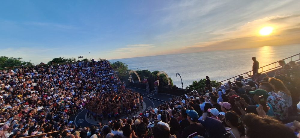 Sejarah Tari Kecak Bali: Pertemuan Timur dan Barat Sejarah Tari Kecak tidak bermula di panggung pertunjukan wisata, melainkan berakar jauh pada ritual keagamaan kuno yang disebut Sanghyang. Sanghyang merupakan tradisi tarian trance yang dilakukan untuk mengusir roh jahat, memohon perlindungan dari dewa, atau menyucikan komunitas dari wabah penyakit. Dalam ritual aslinya, para penari berada dalam kondisi tidak sadar dan diyakini dapat berkomunikasi dengan roh leluhur atau dewa-dewa. Paduan suara pria yang mengiringi ritual ini dikenal sebagai "Cak," yang berfungsi untuk menjaga ritme dan menciptakan atmosfer sakral yang intens. Transformasi dari ritual penyembuhan menuju seni pertunjukan dramatis terjadi pada tahun 1930-an, sebuah periode krusial dalam sejarah seni rupa dan pertunjukan Bali. Inovasi ini merupakan hasil kolaborasi antara I Wayan Limbak, seorang penari dan seniman visioner asal Bali, dengan Walter Spies, seorang pelukis berkebangsaan Jerman yang memiliki keterikatan emosional mendalam dengan budaya Bali. Spies, yang juga memiliki latar belakang dalam dunia musik dan perfilman, melihat potensi artistik yang luar biasa dalam paduan suara Sanghyang. Bersama Limbak, ia merancang sebuah koreografi yang menggabungkan elemen paduan suara "Cak" dengan narasi epos Ramayana. Evolusi ini terdokumentasi dengan baik dalam sejarah perkembangan genre tari Bali. Sebelum tahun 1930-an, elemen "Cak" hanyalah bagian dari ritus pengusiran setan Sanghyang Dedari. Namun, visi Spies dan Limbak di Desa Bona, Kabupaten Gianyar, berhasil mengubah paduan suara tersebut menjadi media penceritaan dramatis. Salah satu katalisator utama transformasi ini adalah keterlibatan Walter Spies dalam pembuatan film Insel der Dämonen (Pulau Iblis) pada tahun 1933 oleh Victor von Plessen. Spies menggunakan medium film untuk mendokumentasikan transformasi genre ini, yang pada gilirannya memperkenalkan estetika Kecak ke audiens internasional. Evolusi dan Pencapaian Historis Tari Kecak Periode/Tahun Milestone Sejarah Keterangan Signifikan Pra-1930 Era Ritual Sanghyang Berfungsi sebagai ritual sakral penyucian dan penolak bala. 1930-an Kolaborasi Limbak-Spies Penciptaan format modern di Desa Bona, Gianyar; integrasi kisah Ramayana. 1933 Dokumentasi Sinematik Penayangan film Insel der Dämonen yang memuat elemen visual Kecak. 1970-an Ekspansi Regional Kecak mulai dipentaskan di luar Gianyar, termasuk di sekolah-sekolah seni di Bali. 1979 Rekor Massal Pertama Pementasan dengan 500 penari pria yang membawakan epos Mahabharata. 2006 Rekor Dunia Tabanan Pementasan kolosal 5.000 penari di Tanah Lot untuk mendongkrak pariwisata. Anatomi Suara: Orkestrasi Gamelan Manusia Keunikan yang paling mencolok dari Tari Kecak adalah absennya instrumen gamelan logam yang biasanya menjadi tulang punggung musik tradisional Bali. Dalam Kecak, instrumen musiknya adalah suara manusia itu sendiri. Orkestrasi vokal ini begitu kompleks sehingga mampu menghasilkan berbagai macam tekstur bunyi, mulai dari dentuman ritmis yang cepat hingga melodi yang melankolis. Paduan suara pria yang duduk melingkar, yang disebut sebagai pengecak, tidak semata-mata meneriakkan kata "cak" secara acak. Terdapat pembagian peran yang sangat teknis di dalam lingkaran tersebut untuk menjaga keharmonisan dan dinamika pertunjukan. Ritme ini dipercaya memiliki frekuensi yang dapat memengaruhi kesadaran, menciptakan suasana magis yang memungkinkan para penari dan penonton terhanyut dalam narasi. Satu-satunya instrumen fisik yang digunakan adalah kincringan, yaitu lonceng kecil yang diikatkan pada kaki para penari lakon (Rama, Sita, Hanuman) untuk memberikan aksen pada setiap langkah dan gerakan tari klasik mereka. Geometri Spiritual: Formasi Lingkaran dan Keseimbangan Rwa Bhineda Visualisasi Tari Kecak dibangun di atas simbolisme yang sarat makna. Formasi para penari yang duduk melingkar bukan sekadar pilihan estetika ruang, melainkan representasi dari kesatuan kosmos dan harmoni alam semesta. Dalam pandangan Hindu Bali, lingkaran melambangkan siklus kehidupan yang abadi, tanpa awal dan tanpa akhir. Posisi ini juga mencerminkan nilai-nilai sosial masyarakat Bali yang menekankan pada gotong royong, kebersamaan, dan kesetaraan; setiap penari berkontribusi pada energi kolektif yang kuat tanpa ada individu yang berusaha menonjolkan diri secara berlebihan. Di tengah lingkaran tersebut, biasanya terdapat damar kecak atau obor api yang berfungsi sebagai titik pusat energi. Api dalam budaya Bali memiliki fungsi ganda: sebagai penerang dan sebagai penyuci (Agni). Menariknya, penggunaan lampu tengah ini awalnya merupakan inovasi teknis yang diperkenalkan oleh Walter Spies untuk kebutuhan pencahayaan saat proses syuting film, agar wajah para penari dapat tertangkap kamera dengan lebih jelas. Namun, seiring berjalannya waktu, elemen ini menjadi bagian permanen dan esensial dalam setiap pertunjukan Kecak, menambah nuansa dramatis dan sakral. Elemen simbolis lainnya adalah kostum kain poleng yang dikenakan oleh para pengecak. Kain dengan motif kotak-kotak hitam dan putih ini merupakan manifestasi dari filosofi Rwa Bhineda, sebuah ajaran fundamental tentang dualisme yang harmonis. Warna hitam melambangkan kegelapan, malam, atau aspek negatif kehidupan, sementara warna putih melambangkan cahaya, siang, atau aspek positif. Dalam pandangan masyarakat Bali, kedua kekuatan ini tidak dapat dipisahkan dan harus selalu berada dalam keseimbangan untuk menjaga keharmonisan dunia. Filosofi ini mengajarkan bahwa kehidupan tidak pernah bersifat absolut; ada keindahan di dalam kegelapan dan ada tantangan di dalam cahaya. Cerita Ramayana dalam Tari Kecak Meskipun instrumen vokalnya berasal dari ritual Sanghyang, jiwa cerita Tari Kecak adalah epos Ramayana. Pertunjukan biasanya menampilkan cuplikan paling dramatis dari perjalanan Pangeran Rama, yang melambangkan pertarungan abadi antara kebajikan (Dharma) dan kejahatan (Adharma). Narasi dimulai di Hutan Dandaka, tempat Rama, istrinya Dewi Sita, dan adiknya Laksmana menjalani masa pengasingan. Di sini, penonton disuguhi drama penculikan yang penuh tipu muslihat. Rahwana, raja raksasa dari Kerajaan Alengka yang dikuasai nafsu serakah, mengirim anak buahnya yang menyamar sebagai kijang emas untuk memikat hati Sita. Ketertarikan Sita pada kijang tersebut memaksa Rama pergi berburu, meninggalkan Sita di bawah perlindungan Laksmana. Adegan yang seringkali menyentuh hati penonton adalah pengorbanan Burung Garuda, Jatayu, yang mencoba mengadang Rahwana saat membawa terbang Sita ke Alengka. Meskipun Jatayu harus gugur setelah sayapnya ditebas oleh kesaktian Rahwana, tindakannya menjadi simbol moral tentang keberanian dan pengorbanan demi membela kehormatan. Puncak ketegangan terjadi saat kemunculan Hanoman, sang kera putih sakti yang menjadi utusan Rama. Hanoman bukan hanya karakter heroik, tetapi juga seringkali menjadi pencair suasana melalui gerakan-gerakannya yang lincah dan jenaka, bahkan terkadang berinteraksi langsung dengan penonton. Adegan "Hanoman Obong" atau pembakaran Hanoman di Alengka adalah momen yang paling ditunggu-tunggu. Di atas bara api yang menyala, penari Hanoman menari dengan tangkas, menyebarkan api ke segala penjuru istana Rahwana, sebuah simbolisasi dari kemurnian yang tidak dapat hangus oleh api kejahatan. Setiap gerakan dalam tarian ini—mulai dari gerakan tangan (mudra), ekspresi wajah (abhinaya), hingga langkah kaki yang anggun—memiliki makna tersendiri dalam teknik tari Bali klasik. Kesetiaan Sita kepada suaminya, keteguhan hati Rama, dan pengabdian Hanoman memberikan pesan moral yang mendalam bagi para penonton: bahwa kebenaran mungkin mengalami hambatan, namun pada akhirnya akan selalu menemukan jalannya untuk menang. So, tokoh-tokoh utama pun hadir: Rama, sang pangeran Sinta, istrinya Hanoman, kera putih yang setia Rahwana, raja raksasa Pertunjukan Tari Kecak biasanya mengangkat kisah Ramayana, yang berfokus pada perjuangan Rama menyelamatkan Sinta dari penculikan Rahwana. Dalam perjalanan tersebut, Rama dibantu oleh Hanoman, tokoh kera putih yang menjadi salah satu karakter paling ikonik dalam pertunjukan ini. Adegan Hanoman bermain dengan api biasanya menjadi bagian paling ditunggu oleh penonton. Yang membuatnya istimewa bukan hanya ceritanya, tetapi cara penyampaiannya. Tanpa dialog panjang, tanpa efek suara modern, semua emosi disampaikan melalui gerakan, ekspresi, dan tentu saja—suara “cak” yang terus mengalun. Pada puncak pertunjukan, adegan api sering kali menjadi klimaks. Api menyala, Hanoman menari di sekitarnya, dan suasana berubah menjadi intens sekaligus memukau. Lokasi Terbaik Menonton Tari Kecak di Bali Berikut beberapa tempat terbaik untuk menyaksikan Tari Kecak Bali: 1. Pura Luhur Uluwatu Lokasi paling populer dengan latar tebing dan sunset langsung ke Samudra Hindia. Suasana sangat dramatis. 2. Pura Dalem Ubud Cocok untuk yang menginap di Ubud. Suasana lebih intim dan kental nuansa budaya. 3. Garuda Wisnu Kencana Area luas dengan fasilitas modern, cocok untuk wisata keluarga. Harga Tiket Tari Kecak Bali Harga tiket menonton Tari Kecak cukup bervariasi tergantung lokasi: Uluwatu: Rp150.000 – Rp200.000 Ubud: Rp100.000 – Rp150.000 GWK: mulai Rp100.000 Harga bisa berubah sewaktu-waktu, terutama saat musim liburan. Jam Pertunjukan Tari Kecak Umumnya, Tari Kecak ditampilkan pada: Pukul 18.00 – 19.00 WITA Durasi: sekitar 60 menit Waktu ini dipilih karena bertepatan dengan sunset, sehingga menciptakan suasana yang lebih dramatis. Betewe, karena pertunjukan ini sangat populer, disarankan untuk datang lebih awal agar mendapatkan tempat duduk terbaik—terutama di lokasi seperti Uluwatu yang sering penuh. DIantara tiga destinasi di atas, saya masih berkunjung yang di Uluwatu. Sore itu, begitu matahari mulai merunduk di cakrawala, semburat jingga keemasan yang membasuh tebing-tebing kapur di Pura Luhur Uluwatu bukan sekadar penanda berakhirnya hari, melainkan aba-aba bagi dimulainya sebuah upacara estetika yang telah memikat dunia selama hampir satu abad. Di sebuah amfiteater terbuka yang bertengger megah di atas Samudra Hindia, ratusan pria duduk melingkar dengan dada telanjang, hanya mengenakan kain poleng hitam-putih yang melilit pinggang mereka. Ketika matahari bersentuhan dengan garis laut, sebuah seruan ritmik memecah keheningan: "cak, cak, cak." Suara itu bukan sekadar iringan musik, melainkan sebuah getaran kolektif yang merambat dari bumi, melalui tubuh-tubuh penari, hingga menyentuh relung spiritual setiap penonton yang hadir. Awalnya pelan. Kemudian semakin kuat, berlapis, dan ritmis. Suara itu seperti gelombang yang datang silih berganti, membentuk harmoni yang tidak biasa. Inilah ciri khas utama Tari Kecak—tarian yang tidak menggunakan alat musik, melainkan mengandalkan vokal kolektif sebagai pengiring utama. Tari Kecak, sebuah mahakarya yang lahir dari rahim tradisi sakral dan visi artistik modern, berdiri sebagai monumen hidup bagi identitas budaya Bali yang tak pernah lekang oleh zaman. Pementasan ini merupakan perpaduan unik antara energi, cerita, dan spiritualitas yang membius. Berbeda dengan tarian Bali lainnya yang mengandalkan dentuman gamelan perunggu, Kecak sepenuhnya mengandalkan instrumen vokal manusia. Harmoni suara yang dihasilkan oleh 50 hingga 150 penari pria ini menciptakan lanskap bunyi yang trance-like, membawa penonton ke dalam dunia epos Ramayana yang penuh dengan pesan moral tentang kesetiaan, keberanian, dan kemenangan kebenaran atas kejahatan. Di balik kemegahan teatrikalnya, Kecak menyimpan lapisan sejarah yang dalam, mulai dari ritual penyucian kuno hingga inovasi seni di tahun 1930-an yang melibatkan seniman lokal dan pelukis mancanegara. Bagi banyak wisatawan, pengalaman ini terasa magis. Suara manusia yang berpadu dalam pola kompleks menciptakan suasana yang hampir transendental. Salah satu alasan mengapa Tari Kecak begitu berkesan adalah lokasi pertunjukannya. Di Pura Luhur Uluwatu, misalnya, panggung terbuka menghadap langsung ke Samudra Hindia. Tebing tinggi menjadi latar alami, sementara matahari terbenam menciptakan pencahayaan dramatis yang sulit ditandingi. Bagi sebagian orang, Tari Kecak mungkin terlihat seperti atraksi wisata. Namun bagi masyarakat Bali, ini adalah bagian dari identitas. Suara “cak” yang berulang bukan sekadar irama, melainkan simbol kebersamaan. Puluhan bahkan ratusan penari bergerak dan bersuara sebagai satu kesatuan—tanpa dominasi individu. Di tengah dunia yang semakin individualistik, pesan ini terasa relevan. Tari Kecak mengajarkan bahwa harmoni tercipta ketika setiap elemen bekerja bersama. Tips Menikmati Tari Kecak Agar pengalaman semakin maksimal, berikut beberapa tips: Datang minimal 30–60 menit sebelum pertunjukan Pilih tempat duduk dengan sudut pandang sunset Gunakan pakaian nyaman (karena duduk cukup lama) Bawa kamera, tapi jangan lupa menikmati momen tanpa layar Dan yang terpenting, datanglah dengan pikiran terbuka. Karena Tari Kecak bukan hanya untuk ditonton, tetapi untuk dirasakan. Harmoni dalam Setiap Suara Saat pertunjukan berakhir dan suara “cak” perlahan mereda, suasana terasa hening. Penonton berdiri, memberikan tepuk tangan panjang, seolah tidak ingin momen itu berakhir. Di langit, matahari telah tenggelam. Namun pengalaman itu tetap tinggal. Tari Kecak adalah bukti bahwa budaya tidak pernah benar-benar diam. Ia hidup, bergerak, dan terus beradaptasi. Di tengah modernitas Bali yang terus berkembang, Tari Kecak tetap menjadi pengingat bahwa akar budaya adalah kekuatan yang menjaga identitas. Tari Kecak bukan sekadar tontonan, melainkan sebuah simfoni kehidupan. Di dalamnya terdengar suara tanah Bali, getaran doa para leluhur, dan semangat kebersamaan yang terus hidup melampaui batas waktu. Ketika suara "cak" terakhir menggema dan api di tengah lingkaran perlahan padam, yang tersisa bukan hanya kenangan akan sebuah pertunjukan, melainkan sebuah pemahaman yang lebih dalam tentang harmoni manusia dengan sesama, alam, dan Tuhan. Bagi siapa pun yang pernah menyaksikannya, Kecak adalah detak jantung Bali yang akan terus berdenyut, mengundang dunia untuk merayakan keindahan dalam kesatuan vokal dan spiritualitas yang tak tergantikan.