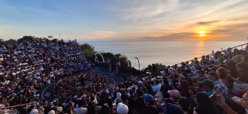 Sejarah Tari Kecak Bali: Pertemuan Timur dan Barat Sejarah Tari Kecak tidak bermula di panggung pertunjukan wisata, melainkan berakar jauh pada ritual keagamaan kuno yang disebut Sanghyang. Sanghyang merupakan tradisi tarian trance yang dilakukan untuk mengusir roh jahat, memohon perlindungan dari dewa, atau menyucikan komunitas dari wabah penyakit. Dalam ritual aslinya, para penari berada dalam kondisi tidak sadar dan diyakini dapat berkomunikasi dengan roh leluhur atau dewa-dewa. Paduan suara pria yang mengiringi ritual ini dikenal sebagai "Cak," yang berfungsi untuk menjaga ritme dan menciptakan atmosfer sakral yang intens. Transformasi dari ritual penyembuhan menuju seni pertunjukan dramatis terjadi pada tahun 1930-an, sebuah periode krusial dalam sejarah seni rupa dan pertunjukan Bali. Inovasi ini merupakan hasil kolaborasi antara I Wayan Limbak, seorang penari dan seniman visioner asal Bali, dengan Walter Spies, seorang pelukis berkebangsaan Jerman yang memiliki keterikatan emosional mendalam dengan budaya Bali. Spies, yang juga memiliki latar belakang dalam dunia musik dan perfilman, melihat potensi artistik yang luar biasa dalam paduan suara Sanghyang. Bersama Limbak, ia merancang sebuah koreografi yang menggabungkan elemen paduan suara "Cak" dengan narasi epos Ramayana. Evolusi ini terdokumentasi dengan baik dalam sejarah perkembangan genre tari Bali. Sebelum tahun 1930-an, elemen "Cak" hanyalah bagian dari ritus pengusiran setan Sanghyang Dedari. Namun, visi Spies dan Limbak di Desa Bona, Kabupaten Gianyar, berhasil mengubah paduan suara tersebut menjadi media penceritaan dramatis. Salah satu katalisator utama transformasi ini adalah keterlibatan Walter Spies dalam pembuatan film Insel der Dämonen (Pulau Iblis) pada tahun 1933 oleh Victor von Plessen. Spies menggunakan medium film untuk mendokumentasikan transformasi genre ini, yang pada gilirannya memperkenalkan estetika Kecak ke audiens internasional. Evolusi dan Pencapaian Historis Tari Kecak Periode/Tahun Milestone Sejarah Keterangan Signifikan Pra-1930 Era Ritual Sanghyang Berfungsi sebagai ritual sakral penyucian dan penolak bala. 1930-an Kolaborasi Limbak-Spies Penciptaan format modern di Desa Bona, Gianyar; integrasi kisah Ramayana. 1933 Dokumentasi Sinematik Penayangan film Insel der Dämonen yang memuat elemen visual Kecak. 1970-an Ekspansi Regional Kecak mulai dipentaskan di luar Gianyar, termasuk di sekolah-sekolah seni di Bali. 1979 Rekor Massal Pertama Pementasan dengan 500 penari pria yang membawakan epos Mahabharata. 2006 Rekor Dunia Tabanan Pementasan kolosal 5.000 penari di Tanah Lot untuk mendongkrak pariwisata. Anatomi Suara: Orkestrasi Gamelan Manusia Keunikan yang paling mencolok dari Tari Kecak adalah absennya instrumen gamelan logam yang biasanya menjadi tulang punggung musik tradisional Bali. Dalam Kecak, instrumen musiknya adalah suara manusia itu sendiri. Orkestrasi vokal ini begitu kompleks sehingga mampu menghasilkan berbagai macam tekstur bunyi, mulai dari dentuman ritmis yang cepat hingga melodi yang melankolis. Paduan suara pria yang duduk melingkar, yang disebut sebagai pengecak, tidak semata-mata meneriakkan kata "cak" secara acak. Terdapat pembagian peran yang sangat teknis di dalam lingkaran tersebut untuk menjaga keharmonisan dan dinamika pertunjukan. Ritme ini dipercaya memiliki frekuensi yang dapat memengaruhi kesadaran, menciptakan suasana magis yang memungkinkan para penari dan penonton terhanyut dalam narasi. Satu-satunya instrumen fisik yang digunakan adalah kincringan, yaitu lonceng kecil yang diikatkan pada kaki para penari lakon (Rama, Sita, Hanuman) untuk memberikan aksen pada setiap langkah dan gerakan tari klasik mereka. Geometri Spiritual: Formasi Lingkaran dan Keseimbangan Rwa Bhineda Visualisasi Tari Kecak dibangun di atas simbolisme yang sarat makna. Formasi para penari yang duduk melingkar bukan sekadar pilihan estetika ruang, melainkan representasi dari kesatuan kosmos dan harmoni alam semesta. Dalam pandangan Hindu Bali, lingkaran melambangkan siklus kehidupan yang abadi, tanpa awal dan tanpa akhir. Posisi ini juga mencerminkan nilai-nilai sosial masyarakat Bali yang menekankan pada gotong royong, kebersamaan, dan kesetaraan; setiap penari berkontribusi pada energi kolektif yang kuat tanpa ada individu yang berusaha menonjolkan diri secara berlebihan. Di tengah lingkaran tersebut, biasanya terdapat damar kecak atau obor api yang berfungsi sebagai titik pusat energi. Api dalam budaya Bali memiliki fungsi ganda: sebagai penerang dan sebagai penyuci (Agni). Menariknya, penggunaan lampu tengah ini awalnya merupakan inovasi teknis yang diperkenalkan oleh Walter Spies untuk kebutuhan pencahayaan saat proses syuting film, agar wajah para penari dapat tertangkap kamera dengan lebih jelas. Namun, seiring berjalannya waktu, elemen ini menjadi bagian permanen dan esensial dalam setiap pertunjukan Kecak, menambah nuansa dramatis dan sakral. Elemen simbolis lainnya adalah kostum kain poleng yang dikenakan oleh para pengecak. Kain dengan motif kotak-kotak hitam dan putih ini merupakan manifestasi dari filosofi Rwa Bhineda, sebuah ajaran fundamental tentang dualisme yang harmonis. Warna hitam melambangkan kegelapan, malam, atau aspek negatif kehidupan, sementara warna putih melambangkan cahaya, siang, atau aspek positif. Dalam pandangan masyarakat Bali, kedua kekuatan ini tidak dapat dipisahkan dan harus selalu berada dalam keseimbangan untuk menjaga keharmonisan dunia. Filosofi ini mengajarkan bahwa kehidupan tidak pernah bersifat absolut; ada keindahan di dalam kegelapan dan ada tantangan di dalam cahaya. Cerita Ramayana dalam Tari Kecak Meskipun instrumen vokalnya berasal dari ritual Sanghyang, jiwa cerita Tari Kecak adalah epos Ramayana. Pertunjukan biasanya menampilkan cuplikan paling dramatis dari perjalanan Pangeran Rama, yang melambangkan pertarungan abadi antara kebajikan (Dharma) dan kejahatan (Adharma). Narasi dimulai di Hutan Dandaka, tempat Rama, istrinya Dewi Sita, dan adiknya Laksmana menjalani masa pengasingan. Di sini, penonton disuguhi drama penculikan yang penuh tipu muslihat. Rahwana, raja raksasa dari Kerajaan Alengka yang dikuasai nafsu serakah, mengirim anak buahnya yang menyamar sebagai kijang emas untuk memikat hati Sita. Ketertarikan Sita pada kijang tersebut memaksa Rama pergi berburu, meninggalkan Sita di bawah perlindungan Laksmana. Adegan yang seringkali menyentuh hati penonton adalah pengorbanan Burung Garuda, Jatayu, yang mencoba mengadang Rahwana saat membawa terbang Sita ke Alengka. Meskipun Jatayu harus gugur setelah sayapnya ditebas oleh kesaktian Rahwana, tindakannya menjadi simbol moral tentang keberanian dan pengorbanan demi membela kehormatan. Puncak ketegangan terjadi saat kemunculan Hanoman, sang kera putih sakti yang menjadi utusan Rama. Hanoman bukan hanya karakter heroik, tetapi juga seringkali menjadi pencair suasana melalui gerakan-gerakannya yang lincah dan jenaka, bahkan terkadang berinteraksi langsung dengan penonton. Adegan "Hanoman Obong" atau pembakaran Hanoman di Alengka adalah momen yang paling ditunggu-tunggu. Di atas bara api yang menyala, penari Hanoman menari dengan tangkas, menyebarkan api ke segala penjuru istana Rahwana, sebuah simbolisasi dari kemurnian yang tidak dapat hangus oleh api kejahatan. Setiap gerakan dalam tarian ini—mulai dari gerakan tangan (mudra), ekspresi wajah (abhinaya), hingga langkah kaki yang anggun—memiliki makna tersendiri dalam teknik tari Bali klasik. Kesetiaan Sita kepada suaminya, keteguhan hati Rama, dan pengabdian Hanoman memberikan pesan moral yang mendalam bagi para penonton: bahwa kebenaran mungkin mengalami hambatan, namun pada akhirnya akan selalu menemukan jalannya untuk menang. So, tokoh-tokoh utama pun hadir: Rama, sang pangeran Sinta, istrinya Hanoman, kera putih yang setia Rahwana, raja raksasa Pertunjukan Tari Kecak biasanya mengangkat kisah Ramayana, yang berfokus pada perjuangan Rama menyelamatkan Sinta dari penculikan Rahwana. Dalam perjalanan tersebut, Rama dibantu oleh Hanoman, tokoh kera putih yang menjadi salah satu karakter paling ikonik dalam pertunjukan ini. Adegan Hanoman bermain dengan api biasanya menjadi bagian paling ditunggu oleh penonton. Yang membuatnya istimewa bukan hanya ceritanya, tetapi cara penyampaiannya. Tanpa dialog panjang, tanpa efek suara modern, semua emosi disampaikan melalui gerakan, ekspresi, dan tentu saja—suara “cak” yang terus mengalun. Pada puncak pertunjukan, adegan api sering kali menjadi klimaks. Api menyala, Hanoman menari di sekitarnya, dan suasana berubah menjadi intens sekaligus memukau. Lokasi Terbaik Menonton Tari Kecak di Bali Berikut beberapa tempat terbaik untuk menyaksikan Tari Kecak Bali: 1. Pura Luhur Uluwatu Lokasi paling populer dengan latar tebing dan sunset langsung ke Samudra Hindia. Suasana sangat dramatis. 2. Pura Dalem Ubud Cocok untuk yang menginap di Ubud. Suasana lebih intim dan kental nuansa budaya. 3. Garuda Wisnu Kencana Area luas dengan fasilitas modern, cocok untuk wisata keluarga. Harga Tiket Tari Kecak Bali Harga tiket menonton Tari Kecak cukup bervariasi tergantung lokasi: Uluwatu: Rp150.000 – Rp200.000 Ubud: Rp100.000 – Rp150.000 GWK: mulai Rp100.000 Harga bisa berubah sewaktu-waktu, terutama saat musim liburan. Jam Pertunjukan Tari Kecak Umumnya, Tari Kecak ditampilkan pada: Pukul 18.00 – 19.00 WITA Durasi: sekitar 60 menit Waktu ini dipilih karena bertepatan dengan sunset, sehingga menciptakan suasana yang lebih dramatis. Betewe, karena pertunjukan ini sangat populer, disarankan untuk datang lebih awal agar mendapatkan tempat duduk terbaik—terutama di lokasi seperti Uluwatu yang sering penuh. DIantara tiga destinasi di atas, saya masih berkunjung yang di Uluwatu. Sore itu, begitu matahari mulai merunduk di cakrawala, semburat jingga keemasan yang membasuh tebing-tebing kapur di Pura Luhur Uluwatu bukan sekadar penanda berakhirnya hari, melainkan aba-aba bagi dimulainya sebuah upacara estetika yang telah memikat dunia selama hampir satu abad. Di sebuah amfiteater terbuka yang bertengger megah di atas Samudra Hindia, ratusan pria duduk melingkar dengan dada telanjang, hanya mengenakan kain poleng hitam-putih yang melilit pinggang mereka. Ketika matahari bersentuhan dengan garis laut, sebuah seruan ritmik memecah keheningan: "cak, cak, cak." Suara itu bukan sekadar iringan musik, melainkan sebuah getaran kolektif yang merambat dari bumi, melalui tubuh-tubuh penari, hingga menyentuh relung spiritual setiap penonton yang hadir. Awalnya pelan. Kemudian semakin kuat, berlapis, dan ritmis. Suara itu seperti gelombang yang datang silih berganti, membentuk harmoni yang tidak biasa. Inilah ciri khas utama Tari Kecak—tarian yang tidak menggunakan alat musik, melainkan mengandalkan vokal kolektif sebagai pengiring utama. Tari Kecak, sebuah mahakarya yang lahir dari rahim tradisi sakral dan visi artistik modern, berdiri sebagai monumen hidup bagi identitas budaya Bali yang tak pernah lekang oleh zaman. Pementasan ini merupakan perpaduan unik antara energi, cerita, dan spiritualitas yang membius. Berbeda dengan tarian Bali lainnya yang mengandalkan dentuman gamelan perunggu, Kecak sepenuhnya mengandalkan instrumen vokal manusia. Harmoni suara yang dihasilkan oleh 50 hingga 150 penari pria ini menciptakan lanskap bunyi yang trance-like, membawa penonton ke dalam dunia epos Ramayana yang penuh dengan pesan moral tentang kesetiaan, keberanian, dan kemenangan kebenaran atas kejahatan. Di balik kemegahan teatrikalnya, Kecak menyimpan lapisan sejarah yang dalam, mulai dari ritual penyucian kuno hingga inovasi seni di tahun 1930-an yang melibatkan seniman lokal dan pelukis mancanegara. Bagi banyak wisatawan, pengalaman ini terasa magis. Suara manusia yang berpadu dalam pola kompleks menciptakan suasana yang hampir transendental. Salah satu alasan mengapa Tari Kecak begitu berkesan adalah lokasi pertunjukannya. Di Pura Luhur Uluwatu, misalnya, panggung terbuka menghadap langsung ke Samudra Hindia. Tebing tinggi menjadi latar alami, sementara matahari terbenam menciptakan pencahayaan dramatis yang sulit ditandingi. Bagi sebagian orang, Tari Kecak mungkin terlihat seperti atraksi wisata. Namun bagi masyarakat Bali, ini adalah bagian dari identitas. Suara “cak” yang berulang bukan sekadar irama, melainkan simbol kebersamaan. Puluhan bahkan ratusan penari bergerak dan bersuara sebagai satu kesatuan—tanpa dominasi individu. Di tengah dunia yang semakin individualistik, pesan ini terasa relevan. Tari Kecak mengajarkan bahwa harmoni tercipta ketika setiap elemen bekerja bersama. Tips Menikmati Tari Kecak Agar pengalaman semakin maksimal, berikut beberapa tips: Datang minimal 30–60 menit sebelum pertunjukan Pilih tempat duduk dengan sudut pandang sunset Gunakan pakaian nyaman (karena duduk cukup lama) Bawa kamera, tapi jangan lupa menikmati momen tanpa layar Dan yang terpenting, datanglah dengan pikiran terbuka. Karena Tari Kecak bukan hanya untuk ditonton, tetapi untuk dirasakan. Harmoni dalam Setiap Suara Saat pertunjukan berakhir dan suara “cak” perlahan mereda, suasana terasa hening. Penonton berdiri, memberikan tepuk tangan panjang, seolah tidak ingin momen itu berakhir. Di langit, matahari telah tenggelam. Namun pengalaman itu tetap tinggal. Tari Kecak adalah bukti bahwa budaya tidak pernah benar-benar diam. Ia hidup, bergerak, dan terus beradaptasi. Di tengah modernitas Bali yang terus berkembang, Tari Kecak tetap menjadi pengingat bahwa akar budaya adalah kekuatan yang menjaga identitas. Tari Kecak bukan sekadar tontonan, melainkan sebuah simfoni kehidupan. Di dalamnya terdengar suara tanah Bali, getaran doa para leluhur, dan semangat kebersamaan yang terus hidup melampaui batas waktu. Ketika suara "cak" terakhir menggema dan api di tengah lingkaran perlahan padam, yang tersisa bukan hanya kenangan akan sebuah pertunjukan, melainkan sebuah pemahaman yang lebih dalam tentang harmoni manusia dengan sesama, alam, dan Tuhan. Bagi siapa pun yang pernah menyaksikannya, Kecak adalah detak jantung Bali yang akan terus berdenyut, mengundang dunia untuk merayakan keindahan dalam kesatuan vokal dan spiritualitas yang tak tergantikan.