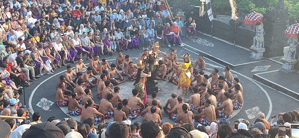 Sejarah Tari Kecak Bali: Pertemuan Timur dan Barat Sejarah Tari Kecak tidak bermula di panggung pertunjukan wisata, melainkan berakar jauh pada ritual keagamaan kuno yang disebut Sanghyang. Sanghyang merupakan tradisi tarian trance yang dilakukan untuk mengusir roh jahat, memohon perlindungan dari dewa, atau menyucikan komunitas dari wabah penyakit. Dalam ritual aslinya, para penari berada dalam kondisi tidak sadar dan diyakini dapat berkomunikasi dengan roh leluhur atau dewa-dewa. Paduan suara pria yang mengiringi ritual ini dikenal sebagai "Cak," yang berfungsi untuk menjaga ritme dan menciptakan atmosfer sakral yang intens. Transformasi dari ritual penyembuhan menuju seni pertunjukan dramatis terjadi pada tahun 1930-an, sebuah periode krusial dalam sejarah seni rupa dan pertunjukan Bali. Inovasi ini merupakan hasil kolaborasi antara I Wayan Limbak, seorang penari dan seniman visioner asal Bali, dengan Walter Spies, seorang pelukis berkebangsaan Jerman yang memiliki keterikatan emosional mendalam dengan budaya Bali. Spies, yang juga memiliki latar belakang dalam dunia musik dan perfilman, melihat potensi artistik yang luar biasa dalam paduan suara Sanghyang. Bersama Limbak, ia merancang sebuah koreografi yang menggabungkan elemen paduan suara "Cak" dengan narasi epos Ramayana. Evolusi ini terdokumentasi dengan baik dalam sejarah perkembangan genre tari Bali. Sebelum tahun 1930-an, elemen "Cak" hanyalah bagian dari ritus pengusiran setan Sanghyang Dedari. Namun, visi Spies dan Limbak di Desa Bona, Kabupaten Gianyar, berhasil mengubah paduan suara tersebut menjadi media penceritaan dramatis. Salah satu katalisator utama transformasi ini adalah keterlibatan Walter Spies dalam pembuatan film Insel der Dämonen (Pulau Iblis) pada tahun 1933 oleh Victor von Plessen. Spies menggunakan medium film untuk mendokumentasikan transformasi genre ini, yang pada gilirannya memperkenalkan estetika Kecak ke audiens internasional. Evolusi dan Pencapaian Historis Tari Kecak Periode/Tahun Milestone Sejarah Keterangan Signifikan Pra-1930 Era Ritual Sanghyang Berfungsi sebagai ritual sakral penyucian dan penolak bala. 1930-an Kolaborasi Limbak-Spies Penciptaan format modern di Desa Bona, Gianyar; integrasi kisah Ramayana. 1933 Dokumentasi Sinematik Penayangan film Insel der Dämonen yang memuat elemen visual Kecak. 1970-an Ekspansi Regional Kecak mulai dipentaskan di luar Gianyar, termasuk di sekolah-sekolah seni di Bali. 1979 Rekor Massal Pertama Pementasan dengan 500 penari pria yang membawakan epos Mahabharata. 2006 Rekor Dunia Tabanan Pementasan kolosal 5.000 penari di Tanah Lot untuk mendongkrak pariwisata. Anatomi Suara: Orkestrasi Gamelan Manusia Keunikan yang paling mencolok dari Tari Kecak adalah absennya instrumen gamelan logam yang biasanya menjadi tulang punggung musik tradisional Bali. Dalam Kecak, instrumen musiknya adalah suara manusia itu sendiri. Orkestrasi vokal ini begitu kompleks sehingga mampu menghasilkan berbagai macam tekstur bunyi, mulai dari dentuman ritmis yang cepat hingga melodi yang melankolis. Paduan suara pria yang duduk melingkar, yang disebut sebagai pengecak, tidak semata-mata meneriakkan kata "cak" secara acak. Terdapat pembagian peran yang sangat teknis di dalam lingkaran tersebut untuk menjaga keharmonisan dan dinamika pertunjukan. Ritme ini dipercaya memiliki frekuensi yang dapat memengaruhi kesadaran, menciptakan suasana magis yang memungkinkan para penari dan penonton terhanyut dalam narasi. Satu-satunya instrumen fisik yang digunakan adalah kincringan, yaitu lonceng kecil yang diikatkan pada kaki para penari lakon (Rama, Sita, Hanuman) untuk memberikan aksen pada setiap langkah dan gerakan tari klasik mereka. Geometri Spiritual: Formasi Lingkaran dan Keseimbangan Rwa Bhineda Visualisasi Tari Kecak dibangun di atas simbolisme yang sarat makna. Formasi para penari yang duduk melingkar bukan sekadar pilihan estetika ruang, melainkan representasi dari kesatuan kosmos dan harmoni alam semesta. Dalam pandangan Hindu Bali, lingkaran melambangkan siklus kehidupan yang abadi, tanpa awal dan tanpa akhir. Posisi ini juga mencerminkan nilai-nilai sosial masyarakat Bali yang menekankan pada gotong royong, kebersamaan, dan kesetaraan; setiap penari berkontribusi pada energi kolektif yang kuat tanpa ada individu yang berusaha menonjolkan diri secara berlebihan. Di tengah lingkaran tersebut, biasanya terdapat damar kecak atau obor api yang berfungsi sebagai titik pusat energi. Api dalam budaya Bali memiliki fungsi ganda: sebagai penerang dan sebagai penyuci (Agni). Menariknya, penggunaan lampu tengah ini awalnya merupakan inovasi teknis yang diperkenalkan oleh Walter Spies untuk kebutuhan pencahayaan saat proses syuting film, agar wajah para penari dapat tertangkap kamera dengan lebih jelas. Namun, seiring berjalannya waktu, elemen ini menjadi bagian permanen dan esensial dalam setiap pertunjukan Kecak, menambah nuansa dramatis dan sakral. Elemen simbolis lainnya adalah kostum kain poleng yang dikenakan oleh para pengecak. Kain dengan motif kotak-kotak hitam dan putih ini merupakan manifestasi dari filosofi Rwa Bhineda, sebuah ajaran fundamental tentang dualisme yang harmonis. Warna hitam melambangkan kegelapan, malam, atau aspek negatif kehidupan, sementara warna putih melambangkan cahaya, siang, atau aspek positif. Dalam pandangan masyarakat Bali, kedua kekuatan ini tidak dapat dipisahkan dan harus selalu berada dalam keseimbangan untuk menjaga keharmonisan dunia. Filosofi ini mengajarkan bahwa kehidupan tidak pernah bersifat absolut; ada keindahan di dalam kegelapan dan ada tantangan di dalam cahaya. Cerita Ramayana dalam Tari Kecak Meskipun instrumen vokalnya berasal dari ritual Sanghyang, jiwa cerita Tari Kecak adalah epos Ramayana. Pertunjukan biasanya menampilkan cuplikan paling dramatis dari perjalanan Pangeran Rama, yang melambangkan pertarungan abadi antara kebajikan (Dharma) dan kejahatan (Adharma). Narasi dimulai di Hutan Dandaka, tempat Rama, istrinya Dewi Sita, dan adiknya Laksmana menjalani masa pengasingan. Di sini, penonton disuguhi drama penculikan yang penuh tipu muslihat. Rahwana, raja raksasa dari Kerajaan Alengka yang dikuasai nafsu serakah, mengirim anak buahnya yang menyamar sebagai kijang emas untuk memikat hati Sita. Ketertarikan Sita pada kijang tersebut memaksa Rama pergi berburu, meninggalkan Sita di bawah perlindungan Laksmana. Adegan yang seringkali menyentuh hati penonton adalah pengorbanan Burung Garuda, Jatayu, yang mencoba mengadang Rahwana saat membawa terbang Sita ke Alengka. Meskipun Jatayu harus gugur setelah sayapnya ditebas oleh kesaktian Rahwana, tindakannya menjadi simbol moral tentang keberanian dan pengorbanan demi membela kehormatan. Puncak ketegangan terjadi saat kemunculan Hanoman, sang kera putih sakti yang menjadi utusan Rama. Hanoman bukan hanya karakter heroik, tetapi juga seringkali menjadi pencair suasana melalui gerakan-gerakannya yang lincah dan jenaka, bahkan terkadang berinteraksi langsung dengan penonton. Adegan "Hanoman Obong" atau pembakaran Hanoman di Alengka adalah momen yang paling ditunggu-tunggu. Di atas bara api yang menyala, penari Hanoman menari dengan tangkas, menyebarkan api ke segala penjuru istana Rahwana, sebuah simbolisasi dari kemurnian yang tidak dapat hangus oleh api kejahatan. Setiap gerakan dalam tarian ini—mulai dari gerakan tangan (mudra), ekspresi wajah (abhinaya), hingga langkah kaki yang anggun—memiliki makna tersendiri dalam teknik tari Bali klasik. Kesetiaan Sita kepada suaminya, keteguhan hati Rama, dan pengabdian Hanoman memberikan pesan moral yang mendalam bagi para penonton: bahwa kebenaran mungkin mengalami hambatan, namun pada akhirnya akan selalu menemukan jalannya untuk menang. So, tokoh-tokoh utama pun hadir: Rama, sang pangeran Sinta, istrinya Hanoman, kera putih yang setia Rahwana, raja raksasa Pertunjukan Tari Kecak biasanya mengangkat kisah Ramayana, yang berfokus pada perjuangan Rama menyelamatkan Sinta dari penculikan Rahwana. Dalam perjalanan tersebut, Rama dibantu oleh Hanoman, tokoh kera putih yang menjadi salah satu karakter paling ikonik dalam pertunjukan ini. Adegan Hanoman bermain dengan api biasanya menjadi bagian paling ditunggu oleh penonton. Yang membuatnya istimewa bukan hanya ceritanya, tetapi cara penyampaiannya. Tanpa dialog panjang, tanpa efek suara modern, semua emosi disampaikan melalui gerakan, ekspresi, dan tentu saja—suara “cak” yang terus mengalun. Pada puncak pertunjukan, adegan api sering kali menjadi klimaks. Api menyala, Hanoman menari di sekitarnya, dan suasana berubah menjadi intens sekaligus memukau. Lokasi Terbaik Menonton Tari Kecak di Bali Berikut beberapa tempat terbaik untuk menyaksikan Tari Kecak Bali: 1. Pura Luhur Uluwatu Lokasi paling populer dengan latar tebing dan sunset langsung ke Samudra Hindia. Suasana sangat dramatis. 2. Pura Dalem Ubud Cocok untuk yang menginap di Ubud. Suasana lebih intim dan kental nuansa budaya. 3. Garuda Wisnu Kencana Area luas dengan fasilitas modern, cocok untuk wisata keluarga. Harga Tiket Tari Kecak Bali Harga tiket menonton Tari Kecak cukup bervariasi tergantung lokasi: Uluwatu: Rp150.000 – Rp200.000 Ubud: Rp100.000 – Rp150.000 GWK: mulai Rp100.000 Harga bisa berubah sewaktu-waktu, terutama saat musim liburan. Jam Pertunjukan Tari Kecak Umumnya, Tari Kecak ditampilkan pada: Pukul 18.00 – 19.00 WITA Durasi: sekitar 60 menit Waktu ini dipilih karena bertepatan dengan sunset, sehingga menciptakan suasana yang lebih dramatis. Betewe, karena pertunjukan ini sangat populer, disarankan untuk datang lebih awal agar mendapatkan tempat duduk terbaik—terutama di lokasi seperti Uluwatu yang sering penuh. DIantara tiga destinasi di atas, saya masih berkunjung yang di Uluwatu. Sore itu, begitu matahari mulai merunduk di cakrawala, semburat jingga keemasan yang membasuh tebing-tebing kapur di Pura Luhur Uluwatu bukan sekadar penanda berakhirnya hari, melainkan aba-aba bagi dimulainya sebuah upacara estetika yang telah memikat dunia selama hampir satu abad. Di sebuah amfiteater terbuka yang bertengger megah di atas Samudra Hindia, ratusan pria duduk melingkar dengan dada telanjang, hanya mengenakan kain poleng hitam-putih yang melilit pinggang mereka. Ketika matahari bersentuhan dengan garis laut, sebuah seruan ritmik memecah keheningan: "cak, cak, cak." Suara itu bukan sekadar iringan musik, melainkan sebuah getaran kolektif yang merambat dari bumi, melalui tubuh-tubuh penari, hingga menyentuh relung spiritual setiap penonton yang hadir. Awalnya pelan. Kemudian semakin kuat, berlapis, dan ritmis. Suara itu seperti gelombang yang datang silih berganti, membentuk harmoni yang tidak biasa. Inilah ciri khas utama Tari Kecak—tarian yang tidak menggunakan alat musik, melainkan mengandalkan vokal kolektif sebagai pengiring utama. Tari Kecak, sebuah mahakarya yang lahir dari rahim tradisi sakral dan visi artistik modern, berdiri sebagai monumen hidup bagi identitas budaya Bali yang tak pernah lekang oleh zaman. Pementasan ini merupakan perpaduan unik antara energi, cerita, dan spiritualitas yang membius. Berbeda dengan tarian Bali lainnya yang mengandalkan dentuman gamelan perunggu, Kecak sepenuhnya mengandalkan instrumen vokal manusia. Harmoni suara yang dihasilkan oleh 50 hingga 150 penari pria ini menciptakan lanskap bunyi yang trance-like, membawa penonton ke dalam dunia epos Ramayana yang penuh dengan pesan moral tentang kesetiaan, keberanian, dan kemenangan kebenaran atas kejahatan. Di balik kemegahan teatrikalnya, Kecak menyimpan lapisan sejarah yang dalam, mulai dari ritual penyucian kuno hingga inovasi seni di tahun 1930-an yang melibatkan seniman lokal dan pelukis mancanegara. Bagi banyak wisatawan, pengalaman ini terasa magis. Suara manusia yang berpadu dalam pola kompleks menciptakan suasana yang hampir transendental. Salah satu alasan mengapa Tari Kecak begitu berkesan adalah lokasi pertunjukannya. Di Pura Luhur Uluwatu, misalnya, panggung terbuka menghadap langsung ke Samudra Hindia. Tebing tinggi menjadi latar alami, sementara matahari terbenam menciptakan pencahayaan dramatis yang sulit ditandingi. Bagi sebagian orang, Tari Kecak mungkin terlihat seperti atraksi wisata. Namun bagi masyarakat Bali, ini adalah bagian dari identitas. Suara “cak” yang berulang bukan sekadar irama, melainkan simbol kebersamaan. Puluhan bahkan ratusan penari bergerak dan bersuara sebagai satu kesatuan—tanpa dominasi individu. Di tengah dunia yang semakin individualistik, pesan ini terasa relevan. Tari Kecak mengajarkan bahwa harmoni tercipta ketika setiap elemen bekerja bersama. Tips Menikmati Tari Kecak Agar pengalaman semakin maksimal, berikut beberapa tips: Datang minimal 30–60 menit sebelum pertunjukan Pilih tempat duduk dengan sudut pandang sunset Gunakan pakaian nyaman (karena duduk cukup lama) Bawa kamera, tapi jangan lupa menikmati momen tanpa layar Dan yang terpenting, datanglah dengan pikiran terbuka. Karena Tari Kecak bukan hanya untuk ditonton, tetapi untuk dirasakan. Harmoni dalam Setiap Suara Saat pertunjukan berakhir dan suara “cak” perlahan mereda, suasana terasa hening. Penonton berdiri, memberikan tepuk tangan panjang, seolah tidak ingin momen itu berakhir. Di langit, matahari telah tenggelam. Namun pengalaman itu tetap tinggal. Tari Kecak adalah bukti bahwa budaya tidak pernah benar-benar diam. Ia hidup, bergerak, dan terus beradaptasi. Di tengah modernitas Bali yang terus berkembang, Tari Kecak tetap menjadi pengingat bahwa akar budaya adalah kekuatan yang menjaga identitas. Tari Kecak bukan sekadar tontonan, melainkan sebuah simfoni kehidupan. Di dalamnya terdengar suara tanah Bali, getaran doa para leluhur, dan semangat kebersamaan yang terus hidup melampaui batas waktu. Ketika suara "cak" terakhir menggema dan api di tengah lingkaran perlahan padam, yang tersisa bukan hanya kenangan akan sebuah pertunjukan, melainkan sebuah pemahaman yang lebih dalam tentang harmoni manusia dengan sesama, alam, dan Tuhan. Bagi siapa pun yang pernah menyaksikannya, Kecak adalah detak jantung Bali yang akan terus berdenyut, mengundang dunia untuk merayakan keindahan dalam kesatuan vokal dan spiritualitas yang tak tergantikan.