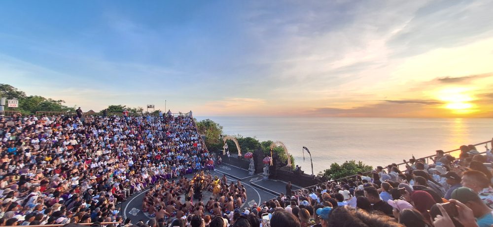 Sejarah Tari Kecak Bali: Pertemuan Timur dan Barat Sejarah Tari Kecak tidak bermula di panggung pertunjukan wisata, melainkan berakar jauh pada ritual keagamaan kuno yang disebut Sanghyang. Sanghyang merupakan tradisi tarian trance yang dilakukan untuk mengusir roh jahat, memohon perlindungan dari dewa, atau menyucikan komunitas dari wabah penyakit. Dalam ritual aslinya, para penari berada dalam kondisi tidak sadar dan diyakini dapat berkomunikasi dengan roh leluhur atau dewa-dewa. Paduan suara pria yang mengiringi ritual ini dikenal sebagai "Cak," yang berfungsi untuk menjaga ritme dan menciptakan atmosfer sakral yang intens. Transformasi dari ritual penyembuhan menuju seni pertunjukan dramatis terjadi pada tahun 1930-an, sebuah periode krusial dalam sejarah seni rupa dan pertunjukan Bali. Inovasi ini merupakan hasil kolaborasi antara I Wayan Limbak, seorang penari dan seniman visioner asal Bali, dengan Walter Spies, seorang pelukis berkebangsaan Jerman yang memiliki keterikatan emosional mendalam dengan budaya Bali. Spies, yang juga memiliki latar belakang dalam dunia musik dan perfilman, melihat potensi artistik yang luar biasa dalam paduan suara Sanghyang. Bersama Limbak, ia merancang sebuah koreografi yang menggabungkan elemen paduan suara "Cak" dengan narasi epos Ramayana. Evolusi ini terdokumentasi dengan baik dalam sejarah perkembangan genre tari Bali. Sebelum tahun 1930-an, elemen "Cak" hanyalah bagian dari ritus pengusiran setan Sanghyang Dedari. Namun, visi Spies dan Limbak di Desa Bona, Kabupaten Gianyar, berhasil mengubah paduan suara tersebut menjadi media penceritaan dramatis. Salah satu katalisator utama transformasi ini adalah keterlibatan Walter Spies dalam pembuatan film Insel der Dämonen (Pulau Iblis) pada tahun 1933 oleh Victor von Plessen. Spies menggunakan medium film untuk mendokumentasikan transformasi genre ini, yang pada gilirannya memperkenalkan estetika Kecak ke audiens internasional. Evolusi dan Pencapaian Historis Tari Kecak Periode/Tahun Milestone Sejarah Keterangan Signifikan Pra-1930 Era Ritual Sanghyang Berfungsi sebagai ritual sakral penyucian dan penolak bala. 1930-an Kolaborasi Limbak-Spies Penciptaan format modern di Desa Bona, Gianyar; integrasi kisah Ramayana. 1933 Dokumentasi Sinematik Penayangan film Insel der Dämonen yang memuat elemen visual Kecak. 1970-an Ekspansi Regional Kecak mulai dipentaskan di luar Gianyar, termasuk di sekolah-sekolah seni di Bali. 1979 Rekor Massal Pertama Pementasan dengan 500 penari pria yang membawakan epos Mahabharata. 2006 Rekor Dunia Tabanan Pementasan kolosal 5.000 penari di Tanah Lot untuk mendongkrak pariwisata. Anatomi Suara: Orkestrasi Gamelan Manusia Keunikan yang paling mencolok dari Tari Kecak adalah absennya instrumen gamelan logam yang biasanya menjadi tulang punggung musik tradisional Bali. Dalam Kecak, instrumen musiknya adalah suara manusia itu sendiri. Orkestrasi vokal ini begitu kompleks sehingga mampu menghasilkan berbagai macam tekstur bunyi, mulai dari dentuman ritmis yang cepat hingga melodi yang melankolis. Paduan suara pria yang duduk melingkar, yang disebut sebagai pengecak, tidak semata-mata meneriakkan kata "cak" secara acak. Terdapat pembagian peran yang sangat teknis di dalam lingkaran tersebut untuk menjaga keharmonisan dan dinamika pertunjukan. Ritme ini dipercaya memiliki frekuensi yang dapat memengaruhi kesadaran, menciptakan suasana magis yang memungkinkan para penari dan penonton terhanyut dalam narasi. Satu-satunya instrumen fisik yang digunakan adalah kincringan, yaitu lonceng kecil yang diikatkan pada kaki para penari lakon (Rama, Sita, Hanuman) untuk memberikan aksen pada setiap langkah dan gerakan tari klasik mereka. Geometri Spiritual: Formasi Lingkaran dan Keseimbangan Rwa Bhineda Visualisasi Tari Kecak dibangun di atas simbolisme yang sarat makna. Formasi para penari yang duduk melingkar bukan sekadar pilihan estetika ruang, melainkan representasi dari kesatuan kosmos dan harmoni alam semesta. Dalam pandangan Hindu Bali, lingkaran melambangkan siklus kehidupan yang abadi, tanpa awal dan tanpa akhir. Posisi ini juga mencerminkan nilai-nilai sosial masyarakat Bali yang menekankan pada gotong royong, kebersamaan, dan kesetaraan; setiap penari berkontribusi pada energi kolektif yang kuat tanpa ada individu yang berusaha menonjolkan diri secara berlebihan. Di tengah lingkaran tersebut, biasanya terdapat damar kecak atau obor api yang berfungsi sebagai titik pusat energi. Api dalam budaya Bali memiliki fungsi ganda: sebagai penerang dan sebagai penyuci (Agni). Menariknya, penggunaan lampu tengah ini awalnya merupakan inovasi teknis yang diperkenalkan oleh Walter Spies untuk kebutuhan pencahayaan saat proses syuting film, agar wajah para penari dapat tertangkap kamera dengan lebih jelas. Namun, seiring berjalannya waktu, elemen ini menjadi bagian permanen dan esensial dalam setiap pertunjukan Kecak, menambah nuansa dramatis dan sakral. Elemen simbolis lainnya adalah kostum kain poleng yang dikenakan oleh para pengecak. Kain dengan motif kotak-kotak hitam dan putih ini merupakan manifestasi dari filosofi Rwa Bhineda, sebuah ajaran fundamental tentang dualisme yang harmonis. Warna hitam melambangkan kegelapan, malam, atau aspek negatif kehidupan, sementara warna putih melambangkan cahaya, siang, atau aspek positif. Dalam pandangan masyarakat Bali, kedua kekuatan ini tidak dapat dipisahkan dan harus selalu berada dalam keseimbangan untuk menjaga keharmonisan dunia. Filosofi ini mengajarkan bahwa kehidupan tidak pernah bersifat absolut; ada keindahan di dalam kegelapan dan ada tantangan di dalam cahaya. Cerita Ramayana dalam Tari Kecak Meskipun instrumen vokalnya berasal dari ritual Sanghyang, jiwa cerita Tari Kecak adalah epos Ramayana. Pertunjukan biasanya menampilkan cuplikan paling dramatis dari perjalanan Pangeran Rama, yang melambangkan pertarungan abadi antara kebajikan (Dharma) dan kejahatan (Adharma). Narasi dimulai di Hutan Dandaka, tempat Rama, istrinya Dewi Sita, dan adiknya Laksmana menjalani masa pengasingan. Di sini, penonton disuguhi drama penculikan yang penuh tipu muslihat. Rahwana, raja raksasa dari Kerajaan Alengka yang dikuasai nafsu serakah, mengirim anak buahnya yang menyamar sebagai kijang emas untuk memikat hati Sita. Ketertarikan Sita pada kijang tersebut memaksa Rama pergi berburu, meninggalkan Sita di bawah perlindungan Laksmana. Adegan yang seringkali menyentuh hati penonton adalah pengorbanan Burung Garuda, Jatayu, yang mencoba mengadang Rahwana saat membawa terbang Sita ke Alengka. Meskipun Jatayu harus gugur setelah sayapnya ditebas oleh kesaktian Rahwana, tindakannya menjadi simbol moral tentang keberanian dan pengorbanan demi membela kehormatan. Puncak ketegangan terjadi saat kemunculan Hanoman, sang kera putih sakti yang menjadi utusan Rama. Hanoman bukan hanya karakter heroik, tetapi juga seringkali menjadi pencair suasana melalui gerakan-gerakannya yang lincah dan jenaka, bahkan terkadang berinteraksi langsung dengan penonton. Adegan "Hanoman Obong" atau pembakaran Hanoman di Alengka adalah momen yang paling ditunggu-tunggu. Di atas bara api yang menyala, penari Hanoman menari dengan tangkas, menyebarkan api ke segala penjuru istana Rahwana, sebuah simbolisasi dari kemurnian yang tidak dapat hangus oleh api kejahatan. Setiap gerakan dalam tarian ini—mulai dari gerakan tangan (mudra), ekspresi wajah (abhinaya), hingga langkah kaki yang anggun—memiliki makna tersendiri dalam teknik tari Bali klasik. Kesetiaan Sita kepada suaminya, keteguhan hati Rama, dan pengabdian Hanoman memberikan pesan moral yang mendalam bagi para penonton: bahwa kebenaran mungkin mengalami hambatan, namun pada akhirnya akan selalu menemukan jalannya untuk menang. So, tokoh-tokoh utama pun hadir: Rama, sang pangeran Sinta, istrinya Hanoman, kera putih yang setia Rahwana, raja raksasa Pertunjukan Tari Kecak biasanya mengangkat kisah Ramayana, yang berfokus pada perjuangan Rama menyelamatkan Sinta dari penculikan Rahwana. Dalam perjalanan tersebut, Rama dibantu oleh Hanoman, tokoh kera putih yang menjadi salah satu karakter paling ikonik dalam pertunjukan ini. Adegan Hanoman bermain dengan api biasanya menjadi bagian paling ditunggu oleh penonton. Yang membuatnya istimewa bukan hanya ceritanya, tetapi cara penyampaiannya. Tanpa dialog panjang, tanpa efek suara modern, semua emosi disampaikan melalui gerakan, ekspresi, dan tentu saja—suara “cak” yang terus mengalun. Pada puncak pertunjukan, adegan api sering kali menjadi klimaks. Api menyala, Hanoman menari di sekitarnya, dan suasana berubah menjadi intens sekaligus memukau. Lokasi Terbaik Menonton Tari Kecak di Bali Berikut beberapa tempat terbaik untuk menyaksikan Tari Kecak Bali: 1. Pura Luhur Uluwatu Lokasi paling populer dengan latar tebing dan sunset langsung ke Samudra Hindia. Suasana sangat dramatis. 2. Pura Dalem Ubud Cocok untuk yang menginap di Ubud. Suasana lebih intim dan kental nuansa budaya. 3. Garuda Wisnu Kencana Area luas dengan fasilitas modern, cocok untuk wisata keluarga. Harga Tiket Tari Kecak Bali Harga tiket menonton Tari Kecak cukup bervariasi tergantung lokasi: Uluwatu: Rp150.000 – Rp200.000 Ubud: Rp100.000 – Rp150.000 GWK: mulai Rp100.000 Harga bisa berubah sewaktu-waktu, terutama saat musim liburan. Jam Pertunjukan Tari Kecak Umumnya, Tari Kecak ditampilkan pada: Pukul 18.00 – 19.00 WITA Durasi: sekitar 60 menit Waktu ini dipilih karena bertepatan dengan sunset, sehingga menciptakan suasana yang lebih dramatis. Betewe, karena pertunjukan ini sangat populer, disarankan untuk datang lebih awal agar mendapatkan tempat duduk terbaik—terutama di lokasi seperti Uluwatu yang sering penuh. DIantara tiga destinasi di atas, saya masih berkunjung yang di Uluwatu. Sore itu, begitu matahari mulai merunduk di cakrawala, semburat jingga keemasan yang membasuh tebing-tebing kapur di Pura Luhur Uluwatu bukan sekadar penanda berakhirnya hari, melainkan aba-aba bagi dimulainya sebuah upacara estetika yang telah memikat dunia selama hampir satu abad. Di sebuah amfiteater terbuka yang bertengger megah di atas Samudra Hindia, ratusan pria duduk melingkar dengan dada telanjang, hanya mengenakan kain poleng hitam-putih yang melilit pinggang mereka. Ketika matahari bersentuhan dengan garis laut, sebuah seruan ritmik memecah keheningan: "cak, cak, cak." Suara itu bukan sekadar iringan musik, melainkan sebuah getaran kolektif yang merambat dari bumi, melalui tubuh-tubuh penari, hingga menyentuh relung spiritual setiap penonton yang hadir. Awalnya pelan. Kemudian semakin kuat, berlapis, dan ritmis. Suara itu seperti gelombang yang datang silih berganti, membentuk harmoni yang tidak biasa. Inilah ciri khas utama Tari Kecak—tarian yang tidak menggunakan alat musik, melainkan mengandalkan vokal kolektif sebagai pengiring utama. Tari Kecak, sebuah mahakarya yang lahir dari rahim tradisi sakral dan visi artistik modern, berdiri sebagai monumen hidup bagi identitas budaya Bali yang tak pernah lekang oleh zaman. Pementasan ini merupakan perpaduan unik antara energi, cerita, dan spiritualitas yang membius. Berbeda dengan tarian Bali lainnya yang mengandalkan dentuman gamelan perunggu, Kecak sepenuhnya mengandalkan instrumen vokal manusia. Harmoni suara yang dihasilkan oleh 50 hingga 150 penari pria ini menciptakan lanskap bunyi yang trance-like, membawa penonton ke dalam dunia epos Ramayana yang penuh dengan pesan moral tentang kesetiaan, keberanian, dan kemenangan kebenaran atas kejahatan. Di balik kemegahan teatrikalnya, Kecak menyimpan lapisan sejarah yang dalam, mulai dari ritual penyucian kuno hingga inovasi seni di tahun 1930-an yang melibatkan seniman lokal dan pelukis mancanegara. Bagi banyak wisatawan, pengalaman ini terasa magis. Suara manusia yang berpadu dalam pola kompleks menciptakan suasana yang hampir transendental. Salah satu alasan mengapa Tari Kecak begitu berkesan adalah lokasi pertunjukannya. Di Pura Luhur Uluwatu, misalnya, panggung terbuka menghadap langsung ke Samudra Hindia. Tebing tinggi menjadi latar alami, sementara matahari terbenam menciptakan pencahayaan dramatis yang sulit ditandingi. Bagi sebagian orang, Tari Kecak mungkin terlihat seperti atraksi wisata. Namun bagi masyarakat Bali, ini adalah bagian dari identitas. Suara “cak” yang berulang bukan sekadar irama, melainkan simbol kebersamaan. Puluhan bahkan ratusan penari bergerak dan bersuara sebagai satu kesatuan—tanpa dominasi individu. Di tengah dunia yang semakin individualistik, pesan ini terasa relevan. Tari Kecak mengajarkan bahwa harmoni tercipta ketika setiap elemen bekerja bersama. Tips Menikmati Tari Kecak Agar pengalaman semakin maksimal, berikut beberapa tips: Datang minimal 30–60 menit sebelum pertunjukan Pilih tempat duduk dengan sudut pandang sunset Gunakan pakaian nyaman (karena duduk cukup lama) Bawa kamera, tapi jangan lupa menikmati momen tanpa layar Dan yang terpenting, datanglah dengan pikiran terbuka. Karena Tari Kecak bukan hanya untuk ditonton, tetapi untuk dirasakan. Harmoni dalam Setiap Suara Saat pertunjukan berakhir dan suara “cak” perlahan mereda, suasana terasa hening. Penonton berdiri, memberikan tepuk tangan panjang, seolah tidak ingin momen itu berakhir. Di langit, matahari telah tenggelam. Namun pengalaman itu tetap tinggal. Tari Kecak adalah bukti bahwa budaya tidak pernah benar-benar diam. Ia hidup, bergerak, dan terus beradaptasi. Di tengah modernitas Bali yang terus berkembang, Tari Kecak tetap menjadi pengingat bahwa akar budaya adalah kekuatan yang menjaga identitas. Tari Kecak bukan sekadar tontonan, melainkan sebuah simfoni kehidupan. Di dalamnya terdengar suara tanah Bali, getaran doa para leluhur, dan semangat kebersamaan yang terus hidup melampaui batas waktu. Ketika suara "cak" terakhir menggema dan api di tengah lingkaran perlahan padam, yang tersisa bukan hanya kenangan akan sebuah pertunjukan, melainkan sebuah pemahaman yang lebih dalam tentang harmoni manusia dengan sesama, alam, dan Tuhan. Bagi siapa pun yang pernah menyaksikannya, Kecak adalah detak jantung Bali yang akan terus berdenyut, mengundang dunia untuk merayakan keindahan dalam kesatuan vokal dan spiritualitas yang tak tergantikan.