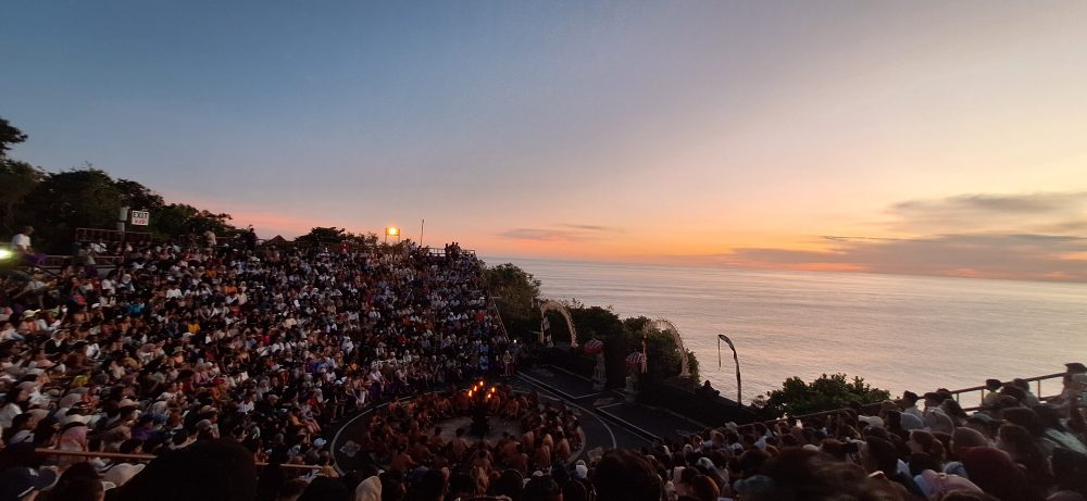 Sejarah Tari Kecak Bali: Pertemuan Timur dan Barat Sejarah Tari Kecak tidak bermula di panggung pertunjukan wisata, melainkan berakar jauh pada ritual keagamaan kuno yang disebut Sanghyang. Sanghyang merupakan tradisi tarian trance yang dilakukan untuk mengusir roh jahat, memohon perlindungan dari dewa, atau menyucikan komunitas dari wabah penyakit. Dalam ritual aslinya, para penari berada dalam kondisi tidak sadar dan diyakini dapat berkomunikasi dengan roh leluhur atau dewa-dewa. Paduan suara pria yang mengiringi ritual ini dikenal sebagai "Cak," yang berfungsi untuk menjaga ritme dan menciptakan atmosfer sakral yang intens. Transformasi dari ritual penyembuhan menuju seni pertunjukan dramatis terjadi pada tahun 1930-an, sebuah periode krusial dalam sejarah seni rupa dan pertunjukan Bali. Inovasi ini merupakan hasil kolaborasi antara I Wayan Limbak, seorang penari dan seniman visioner asal Bali, dengan Walter Spies, seorang pelukis berkebangsaan Jerman yang memiliki keterikatan emosional mendalam dengan budaya Bali. Spies, yang juga memiliki latar belakang dalam dunia musik dan perfilman, melihat potensi artistik yang luar biasa dalam paduan suara Sanghyang. Bersama Limbak, ia merancang sebuah koreografi yang menggabungkan elemen paduan suara "Cak" dengan narasi epos Ramayana. Evolusi ini terdokumentasi dengan baik dalam sejarah perkembangan genre tari Bali. Sebelum tahun 1930-an, elemen "Cak" hanyalah bagian dari ritus pengusiran setan Sanghyang Dedari. Namun, visi Spies dan Limbak di Desa Bona, Kabupaten Gianyar, berhasil mengubah paduan suara tersebut menjadi media penceritaan dramatis. Salah satu katalisator utama transformasi ini adalah keterlibatan Walter Spies dalam pembuatan film Insel der Dämonen (Pulau Iblis) pada tahun 1933 oleh Victor von Plessen. Spies menggunakan medium film untuk mendokumentasikan transformasi genre ini, yang pada gilirannya memperkenalkan estetika Kecak ke audiens internasional. Evolusi dan Pencapaian Historis Tari Kecak Periode/Tahun Milestone Sejarah Keterangan Signifikan Pra-1930 Era Ritual Sanghyang Berfungsi sebagai ritual sakral penyucian dan penolak bala. 1930-an Kolaborasi Limbak-Spies Penciptaan format modern di Desa Bona, Gianyar; integrasi kisah Ramayana. 1933 Dokumentasi Sinematik Penayangan film Insel der Dämonen yang memuat elemen visual Kecak. 1970-an Ekspansi Regional Kecak mulai dipentaskan di luar Gianyar, termasuk di sekolah-sekolah seni di Bali. 1979 Rekor Massal Pertama Pementasan dengan 500 penari pria yang membawakan epos Mahabharata. 2006 Rekor Dunia Tabanan Pementasan kolosal 5.000 penari di Tanah Lot untuk mendongkrak pariwisata. Anatomi Suara: Orkestrasi Gamelan Manusia Keunikan yang paling mencolok dari Tari Kecak adalah absennya instrumen gamelan logam yang biasanya menjadi tulang punggung musik tradisional Bali. Dalam Kecak, instrumen musiknya adalah suara manusia itu sendiri. Orkestrasi vokal ini begitu kompleks sehingga mampu menghasilkan berbagai macam tekstur bunyi, mulai dari dentuman ritmis yang cepat hingga melodi yang melankolis. Paduan suara pria yang duduk melingkar, yang disebut sebagai pengecak, tidak semata-mata meneriakkan kata "cak" secara acak. Terdapat pembagian peran yang sangat teknis di dalam lingkaran tersebut untuk menjaga keharmonisan dan dinamika pertunjukan. Ritme ini dipercaya memiliki frekuensi yang dapat memengaruhi kesadaran, menciptakan suasana magis yang memungkinkan para penari dan penonton terhanyut dalam narasi. Satu-satunya instrumen fisik yang digunakan adalah kincringan, yaitu lonceng kecil yang diikatkan pada kaki para penari lakon (Rama, Sita, Hanuman) untuk memberikan aksen pada setiap langkah dan gerakan tari klasik mereka. Geometri Spiritual: Formasi Lingkaran dan Keseimbangan Rwa Bhineda Visualisasi Tari Kecak dibangun di atas simbolisme yang sarat makna. Formasi para penari yang duduk melingkar bukan sekadar pilihan estetika ruang, melainkan representasi dari kesatuan kosmos dan harmoni alam semesta. Dalam pandangan Hindu Bali, lingkaran melambangkan siklus kehidupan yang abadi, tanpa awal dan tanpa akhir. Posisi ini juga mencerminkan nilai-nilai sosial masyarakat Bali yang menekankan pada gotong royong, kebersamaan, dan kesetaraan; setiap penari berkontribusi pada energi kolektif yang kuat tanpa ada individu yang berusaha menonjolkan diri secara berlebihan. Di tengah lingkaran tersebut, biasanya terdapat damar kecak atau obor api yang berfungsi sebagai titik pusat energi. Api dalam budaya Bali memiliki fungsi ganda: sebagai penerang dan sebagai penyuci (Agni). Menariknya, penggunaan lampu tengah ini awalnya merupakan inovasi teknis yang diperkenalkan oleh Walter Spies untuk kebutuhan pencahayaan saat proses syuting film, agar wajah para penari dapat tertangkap kamera dengan lebih jelas. Namun, seiring berjalannya waktu, elemen ini menjadi bagian permanen dan esensial dalam setiap pertunjukan Kecak, menambah nuansa dramatis dan sakral. Elemen simbolis lainnya adalah kostum kain poleng yang dikenakan oleh para pengecak. Kain dengan motif kotak-kotak hitam dan putih ini merupakan manifestasi dari filosofi Rwa Bhineda, sebuah ajaran fundamental tentang dualisme yang harmonis. Warna hitam melambangkan kegelapan, malam, atau aspek negatif kehidupan, sementara warna putih melambangkan cahaya, siang, atau aspek positif. Dalam pandangan masyarakat Bali, kedua kekuatan ini tidak dapat dipisahkan dan harus selalu berada dalam keseimbangan untuk menjaga keharmonisan dunia. Filosofi ini mengajarkan bahwa kehidupan tidak pernah bersifat absolut; ada keindahan di dalam kegelapan dan ada tantangan di dalam cahaya. Cerita Ramayana dalam Tari Kecak Meskipun instrumen vokalnya berasal dari ritual Sanghyang, jiwa cerita Tari Kecak adalah epos Ramayana. Pertunjukan biasanya menampilkan cuplikan paling dramatis dari perjalanan Pangeran Rama, yang melambangkan pertarungan abadi antara kebajikan (Dharma) dan kejahatan (Adharma). Narasi dimulai di Hutan Dandaka, tempat Rama, istrinya Dewi Sita, dan adiknya Laksmana menjalani masa pengasingan. Di sini, penonton disuguhi drama penculikan yang penuh tipu muslihat. Rahwana, raja raksasa dari Kerajaan Alengka yang dikuasai nafsu serakah, mengirim anak buahnya yang menyamar sebagai kijang emas untuk memikat hati Sita. Ketertarikan Sita pada kijang tersebut memaksa Rama pergi berburu, meninggalkan Sita di bawah perlindungan Laksmana. Adegan yang seringkali menyentuh hati penonton adalah pengorbanan Burung Garuda, Jatayu, yang mencoba mengadang Rahwana saat membawa terbang Sita ke Alengka. Meskipun Jatayu harus gugur setelah sayapnya ditebas oleh kesaktian Rahwana, tindakannya menjadi simbol moral tentang keberanian dan pengorbanan demi membela kehormatan. Puncak ketegangan terjadi saat kemunculan Hanoman, sang kera putih sakti yang menjadi utusan Rama. Hanoman bukan hanya karakter heroik, tetapi juga seringkali menjadi pencair suasana melalui gerakan-gerakannya yang lincah dan jenaka, bahkan terkadang berinteraksi langsung dengan penonton. Adegan "Hanoman Obong" atau pembakaran Hanoman di Alengka adalah momen yang paling ditunggu-tunggu. Di atas bara api yang menyala, penari Hanoman menari dengan tangkas, menyebarkan api ke segala penjuru istana Rahwana, sebuah simbolisasi dari kemurnian yang tidak dapat hangus oleh api kejahatan. Setiap gerakan dalam tarian ini—mulai dari gerakan tangan (mudra), ekspresi wajah (abhinaya), hingga langkah kaki yang anggun—memiliki makna tersendiri dalam teknik tari Bali klasik. Kesetiaan Sita kepada suaminya, keteguhan hati Rama, dan pengabdian Hanoman memberikan pesan moral yang mendalam bagi para penonton: bahwa kebenaran mungkin mengalami hambatan, namun pada akhirnya akan selalu menemukan jalannya untuk menang. So, tokoh-tokoh utama pun hadir: Rama, sang pangeran Sinta, istrinya Hanoman, kera putih yang setia Rahwana, raja raksasa Pertunjukan Tari Kecak biasanya mengangkat kisah Ramayana, yang berfokus pada perjuangan Rama menyelamatkan Sinta dari penculikan Rahwana. Dalam perjalanan tersebut, Rama dibantu oleh Hanoman, tokoh kera putih yang menjadi salah satu karakter paling ikonik dalam pertunjukan ini. Adegan Hanoman bermain dengan api biasanya menjadi bagian paling ditunggu oleh penonton. Yang membuatnya istimewa bukan hanya ceritanya, tetapi cara penyampaiannya. Tanpa dialog panjang, tanpa efek suara modern, semua emosi disampaikan melalui gerakan, ekspresi, dan tentu saja—suara “cak” yang terus mengalun. Pada puncak pertunjukan, adegan api sering kali menjadi klimaks. Api menyala, Hanoman menari di sekitarnya, dan suasana berubah menjadi intens sekaligus memukau. Lokasi Terbaik Menonton Tari Kecak di Bali Berikut beberapa tempat terbaik untuk menyaksikan Tari Kecak Bali: 1. Pura Luhur Uluwatu Lokasi paling populer dengan latar tebing dan sunset langsung ke Samudra Hindia. Suasana sangat dramatis. 2. Pura Dalem Ubud Cocok untuk yang menginap di Ubud. Suasana lebih intim dan kental nuansa budaya. 3. Garuda Wisnu Kencana Area luas dengan fasilitas modern, cocok untuk wisata keluarga. Harga Tiket Tari Kecak Bali Harga tiket menonton Tari Kecak cukup bervariasi tergantung lokasi: Uluwatu: Rp150.000 – Rp200.000 Ubud: Rp100.000 – Rp150.000 GWK: mulai Rp100.000 Harga bisa berubah sewaktu-waktu, terutama saat musim liburan. Jam Pertunjukan Tari Kecak Umumnya, Tari Kecak ditampilkan pada: Pukul 18.00 – 19.00 WITA Durasi: sekitar 60 menit Waktu ini dipilih karena bertepatan dengan sunset, sehingga menciptakan suasana yang lebih dramatis. Betewe, karena pertunjukan ini sangat populer, disarankan untuk datang lebih awal agar mendapatkan tempat duduk terbaik—terutama di lokasi seperti Uluwatu yang sering penuh. DIantara tiga destinasi di atas, saya masih berkunjung yang di Uluwatu. Sore itu, begitu matahari mulai merunduk di cakrawala, semburat jingga keemasan yang membasuh tebing-tebing kapur di Pura Luhur Uluwatu bukan sekadar penanda berakhirnya hari, melainkan aba-aba bagi dimulainya sebuah upacara estetika yang telah memikat dunia selama hampir satu abad. Di sebuah amfiteater terbuka yang bertengger megah di atas Samudra Hindia, ratusan pria duduk melingkar dengan dada telanjang, hanya mengenakan kain poleng hitam-putih yang melilit pinggang mereka. Ketika matahari bersentuhan dengan garis laut, sebuah seruan ritmik memecah keheningan: "cak, cak, cak." Suara itu bukan sekadar iringan musik, melainkan sebuah getaran kolektif yang merambat dari bumi, melalui tubuh-tubuh penari, hingga menyentuh relung spiritual setiap penonton yang hadir. Awalnya pelan. Kemudian semakin kuat, berlapis, dan ritmis. Suara itu seperti gelombang yang datang silih berganti, membentuk harmoni yang tidak biasa. Inilah ciri khas utama Tari Kecak—tarian yang tidak menggunakan alat musik, melainkan mengandalkan vokal kolektif sebagai pengiring utama. Tari Kecak, sebuah mahakarya yang lahir dari rahim tradisi sakral dan visi artistik modern, berdiri sebagai monumen hidup bagi identitas budaya Bali yang tak pernah lekang oleh zaman. Pementasan ini merupakan perpaduan unik antara energi, cerita, dan spiritualitas yang membius. Berbeda dengan tarian Bali lainnya yang mengandalkan dentuman gamelan perunggu, Kecak sepenuhnya mengandalkan instrumen vokal manusia. Harmoni suara yang dihasilkan oleh 50 hingga 150 penari pria ini menciptakan lanskap bunyi yang trance-like, membawa penonton ke dalam dunia epos Ramayana yang penuh dengan pesan moral tentang kesetiaan, keberanian, dan kemenangan kebenaran atas kejahatan. Di balik kemegahan teatrikalnya, Kecak menyimpan lapisan sejarah yang dalam, mulai dari ritual penyucian kuno hingga inovasi seni di tahun 1930-an yang melibatkan seniman lokal dan pelukis mancanegara. Bagi banyak wisatawan, pengalaman ini terasa magis. Suara manusia yang berpadu dalam pola kompleks menciptakan suasana yang hampir transendental. Salah satu alasan mengapa Tari Kecak begitu berkesan adalah lokasi pertunjukannya. Di Pura Luhur Uluwatu, misalnya, panggung terbuka menghadap langsung ke Samudra Hindia. Tebing tinggi menjadi latar alami, sementara matahari terbenam menciptakan pencahayaan dramatis yang sulit ditandingi. Bagi sebagian orang, Tari Kecak mungkin terlihat seperti atraksi wisata. Namun bagi masyarakat Bali, ini adalah bagian dari identitas. Suara “cak” yang berulang bukan sekadar irama, melainkan simbol kebersamaan. Puluhan bahkan ratusan penari bergerak dan bersuara sebagai satu kesatuan—tanpa dominasi individu. Di tengah dunia yang semakin individualistik, pesan ini terasa relevan. Tari Kecak mengajarkan bahwa harmoni tercipta ketika setiap elemen bekerja bersama. Tips Menikmati Tari Kecak Agar pengalaman semakin maksimal, berikut beberapa tips: Datang minimal 30–60 menit sebelum pertunjukan Pilih tempat duduk dengan sudut pandang sunset Gunakan pakaian nyaman (karena duduk cukup lama) Bawa kamera, tapi jangan lupa menikmati momen tanpa layar Dan yang terpenting, datanglah dengan pikiran terbuka. Karena Tari Kecak bukan hanya untuk ditonton, tetapi untuk dirasakan. Harmoni dalam Setiap Suara Saat pertunjukan berakhir dan suara “cak” perlahan mereda, suasana terasa hening. Penonton berdiri, memberikan tepuk tangan panjang, seolah tidak ingin momen itu berakhir. Di langit, matahari telah tenggelam. Namun pengalaman itu tetap tinggal. Tari Kecak adalah bukti bahwa budaya tidak pernah benar-benar diam. Ia hidup, bergerak, dan terus beradaptasi. Di tengah modernitas Bali yang terus berkembang, Tari Kecak tetap menjadi pengingat bahwa akar budaya adalah kekuatan yang menjaga identitas. Tari Kecak bukan sekadar tontonan, melainkan sebuah simfoni kehidupan. Di dalamnya terdengar suara tanah Bali, getaran doa para leluhur, dan semangat kebersamaan yang terus hidup melampaui batas waktu. Ketika suara "cak" terakhir menggema dan api di tengah lingkaran perlahan padam, yang tersisa bukan hanya kenangan akan sebuah pertunjukan, melainkan sebuah pemahaman yang lebih dalam tentang harmoni manusia dengan sesama, alam, dan Tuhan. Bagi siapa pun yang pernah menyaksikannya, Kecak adalah detak jantung Bali yang akan terus berdenyut, mengundang dunia untuk merayakan keindahan dalam kesatuan vokal dan spiritualitas yang tak tergantikan.